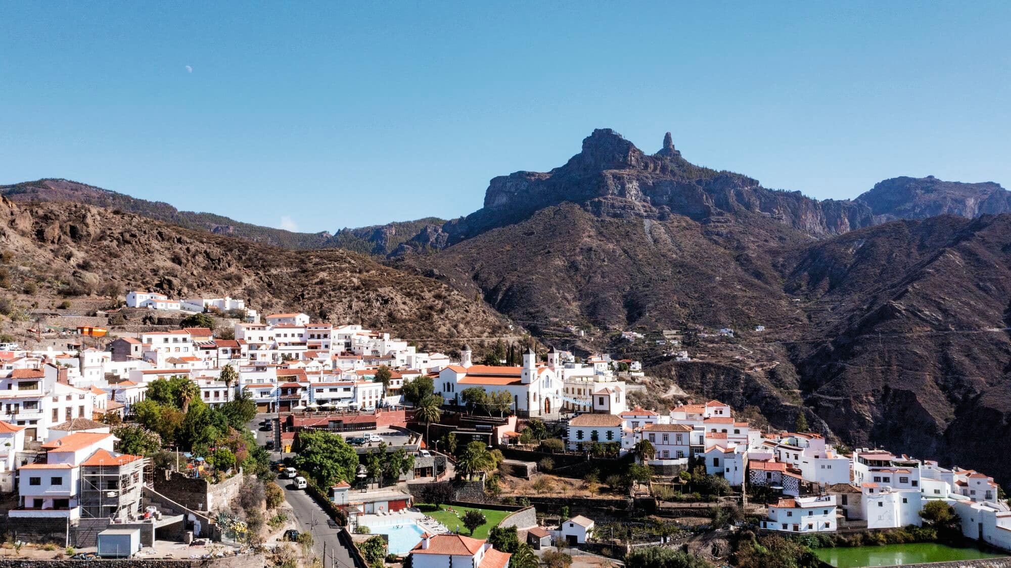 A traditional white-walled Spanish village with terracotta roofs nestled at the base of dramatic rocky mountains under a clear blue sky.