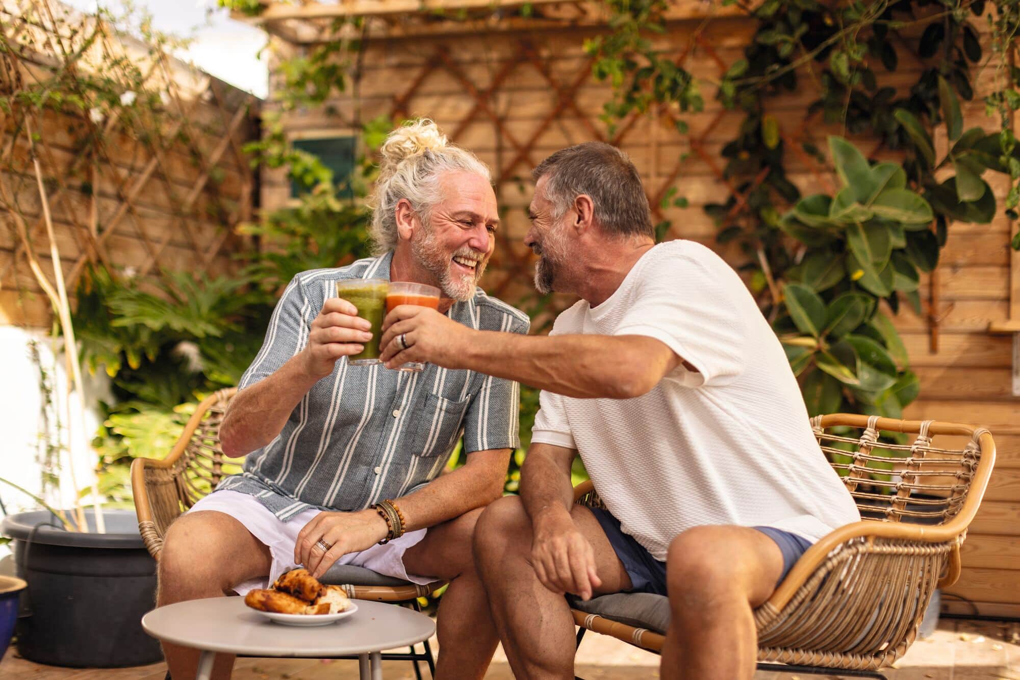 Two people sitting in wicker chairs on an outdoor patio, smiling and holding cups while enjoying breakfast pastries on a round table.