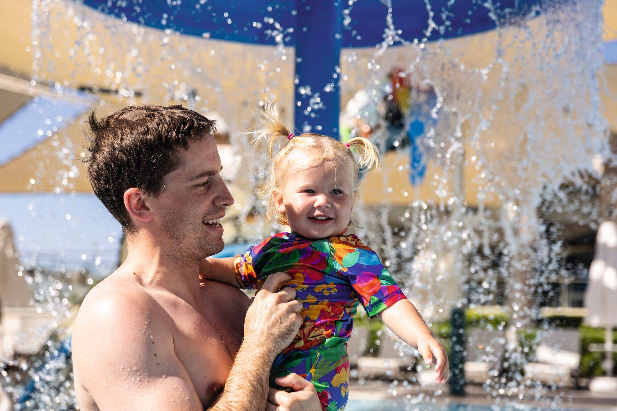 Smiling father holding happy toddler with blonde pigtails in colourful swimsuit at water park splash pad with fountains.