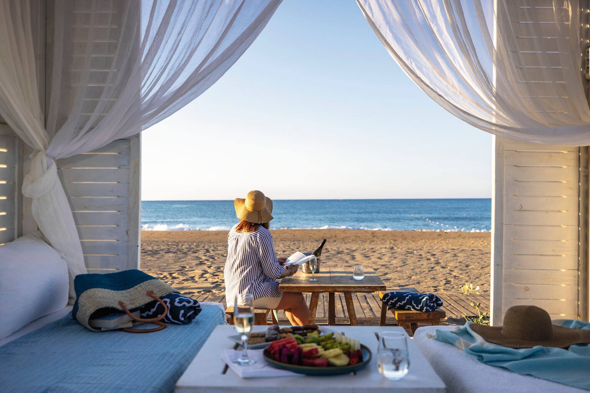 Woman in striped shirt and sun hat relaxing on wooden deck of white beach cabana, with champagne and fruit in foreground.