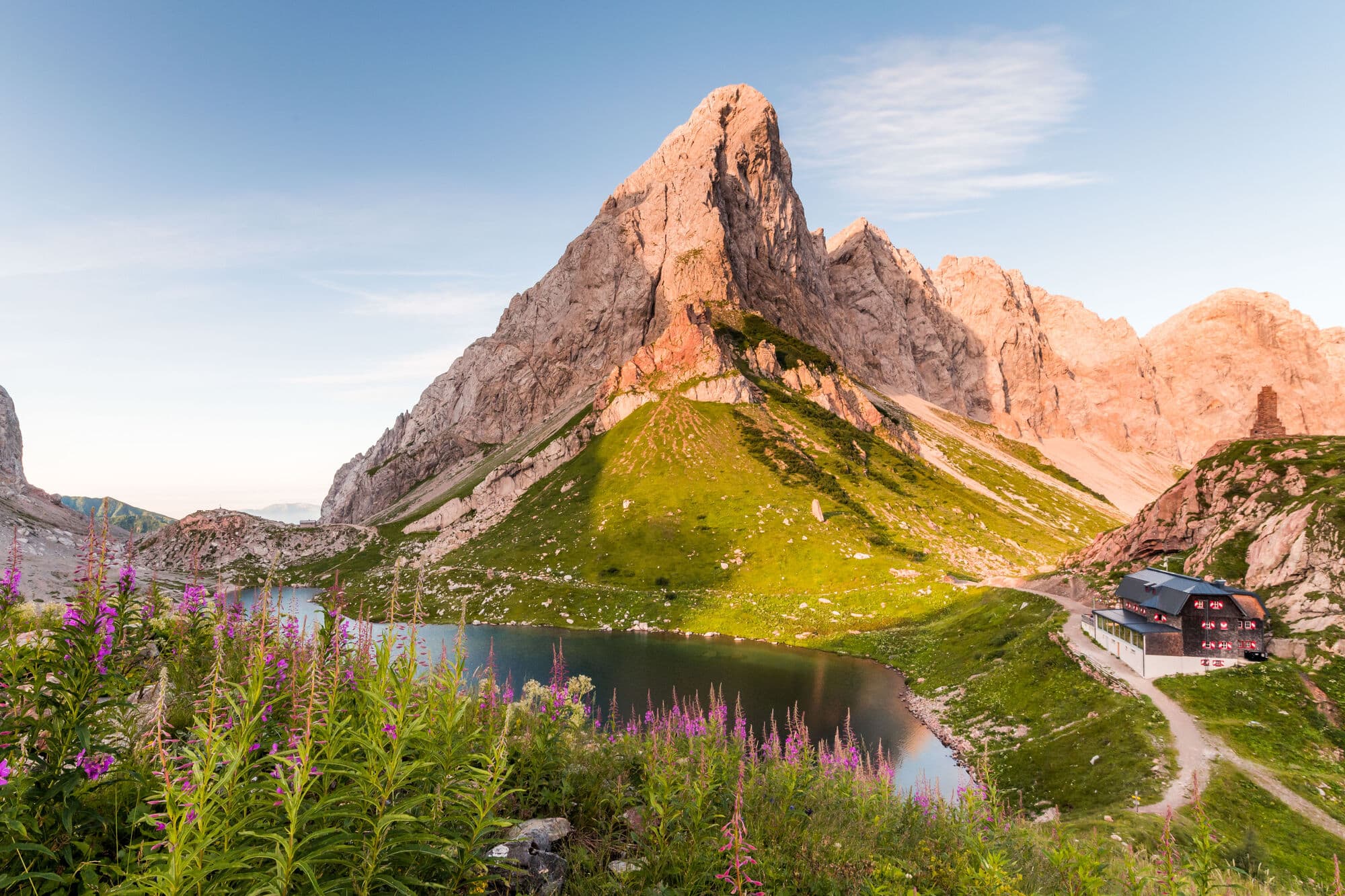 A pointy mountain peak surrounded by lush greenery, a stream and purple wild flowers.
