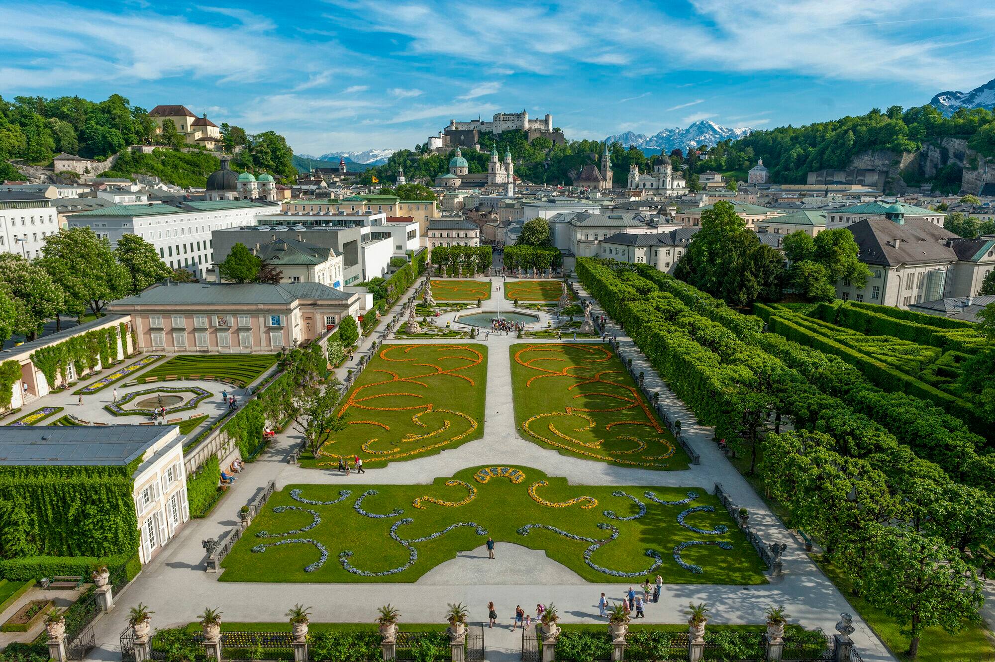 Panoramic view of Salzburg with its manicured gardens in the foreground, historic cityscape featuring the Hohensalzburg Fortress, and snow-capped Alpine mountains creating a stunning backdrop.