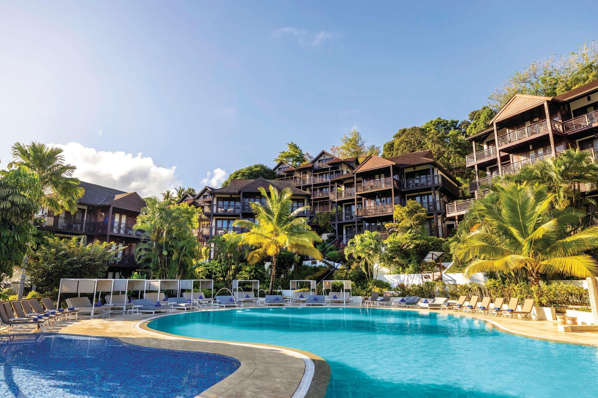 Resort view with two swimming pools in foreground—one deep blue, one turquoise surrounded by white sun loungers, tropical palm trees, and multi-story dark brown buildings.