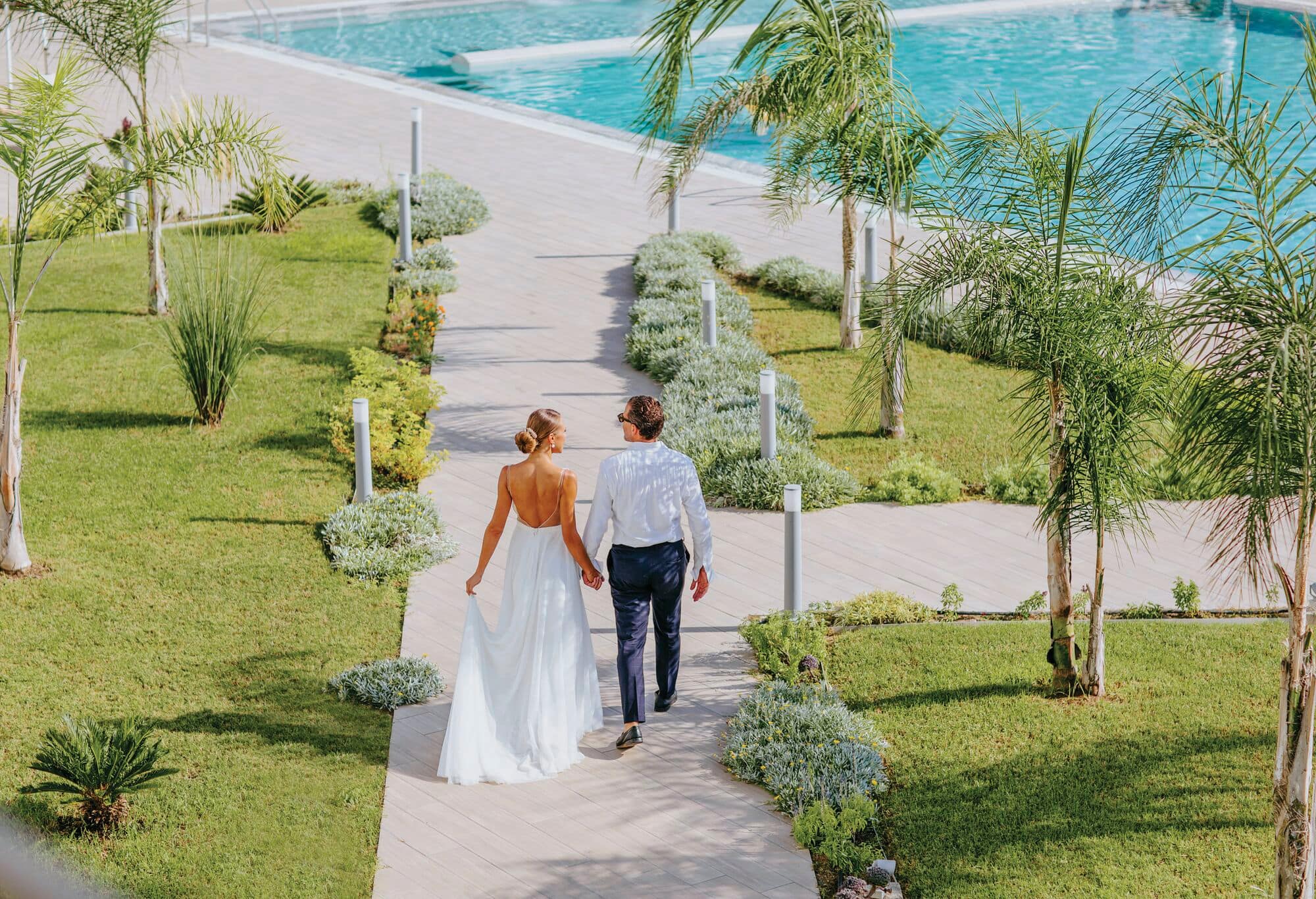 A bride is escorted through a hotel garden. Miniature palms and a vibrant blue pool betray the tropical setting.