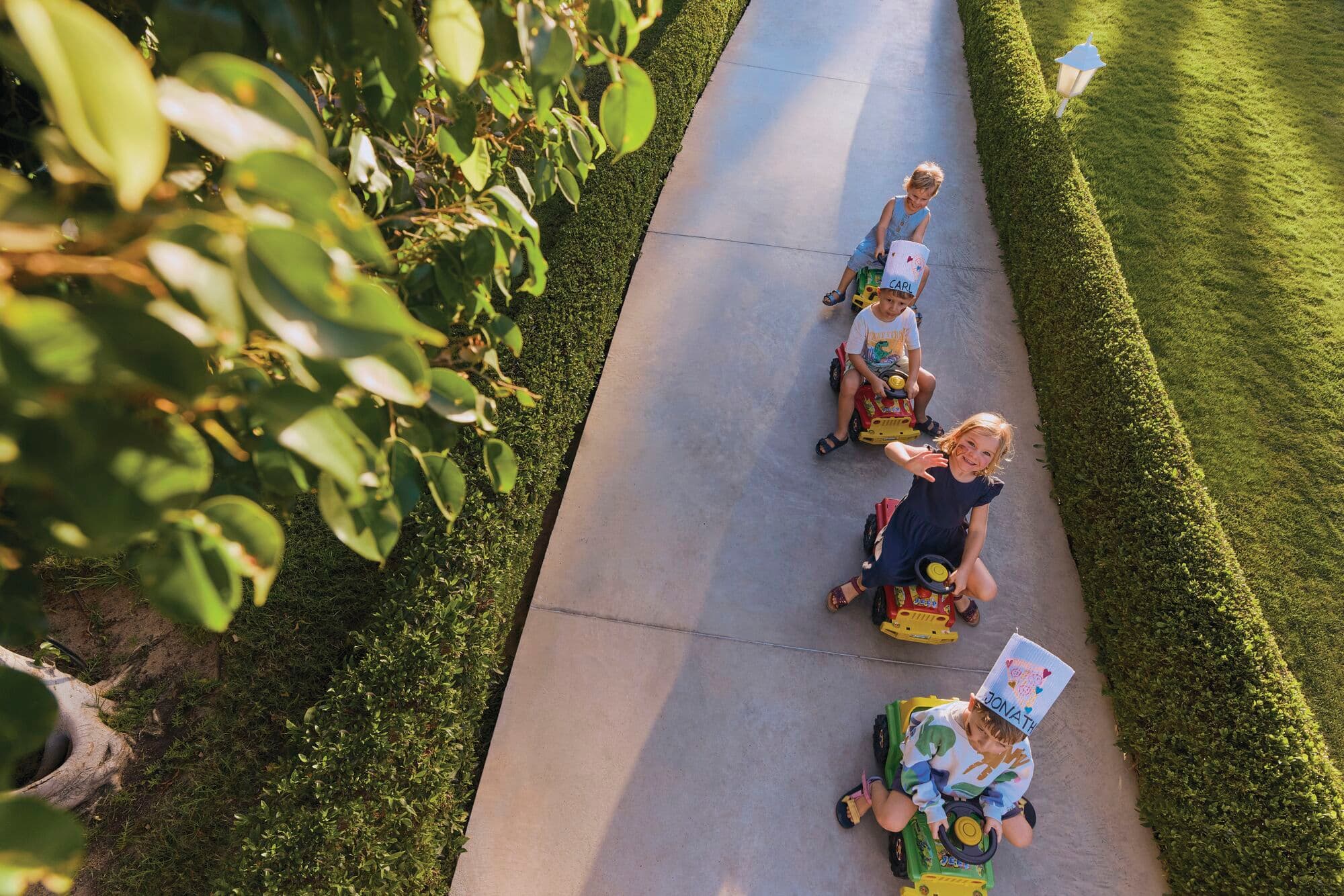 Aerial view of children riding yellow toy carts in a line along a concrete garden path.