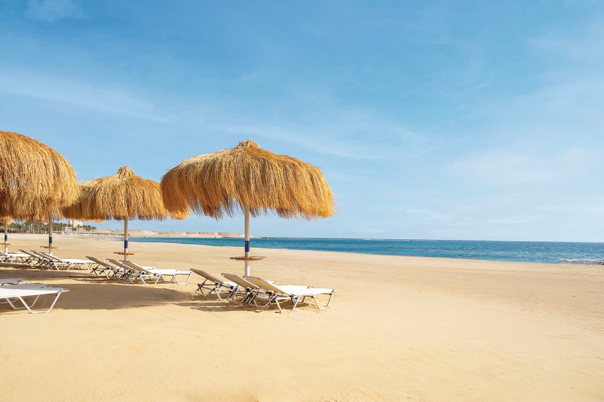 Sunloungers on a beach look out to sea under golden grass umbrellas.