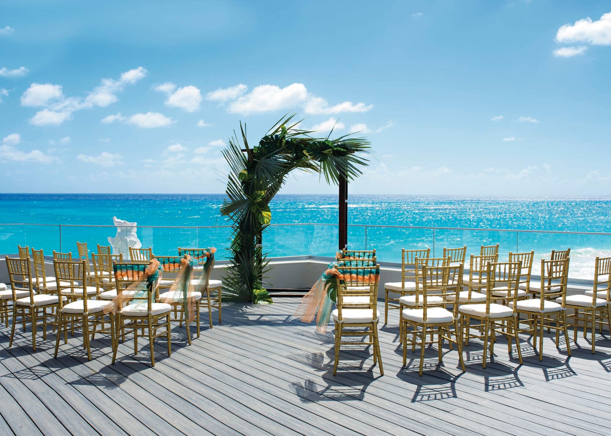 An outdoor wedding set-up on a wooden deck overlooking the sea. Rows of gold chairs are arranged on both sides of an aisle leading to a an arch decorated with palm leaves.