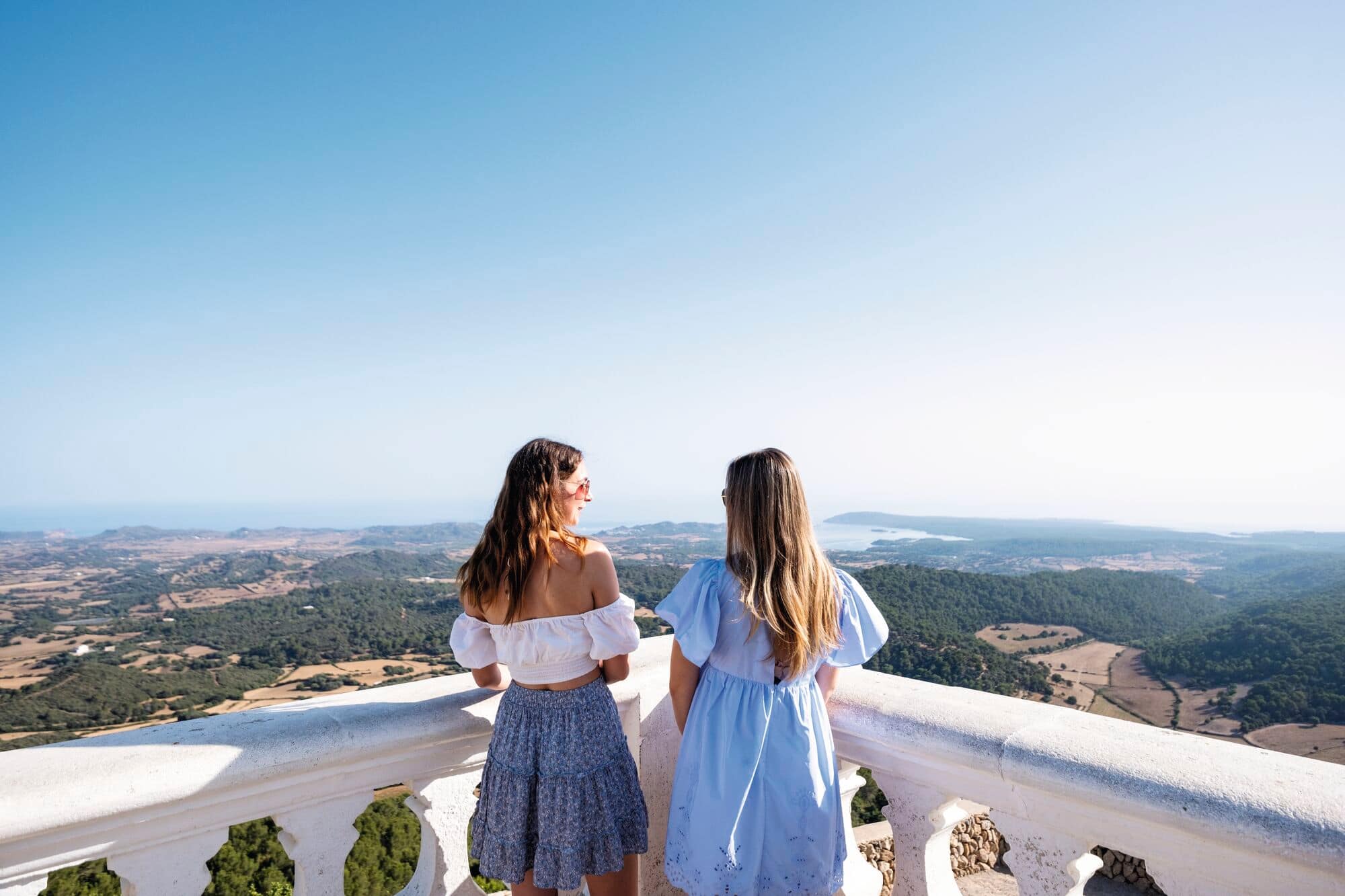 Two women stand at a white terrace balustrade, viewed from behind, overlooking a vast Mediterranean landscape.