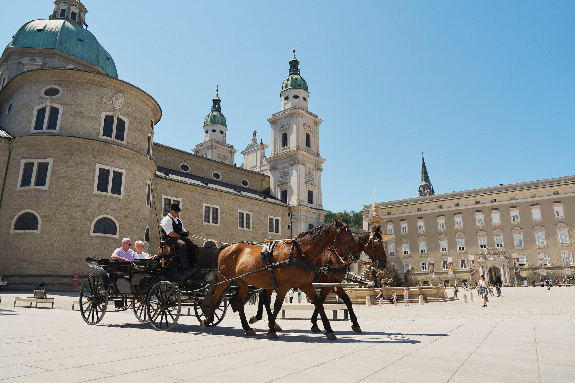 Horse-drawn carriage with tourists in Salzburg's historic square, surrounded by baroque architecture with a domed cathedral under blue skies.