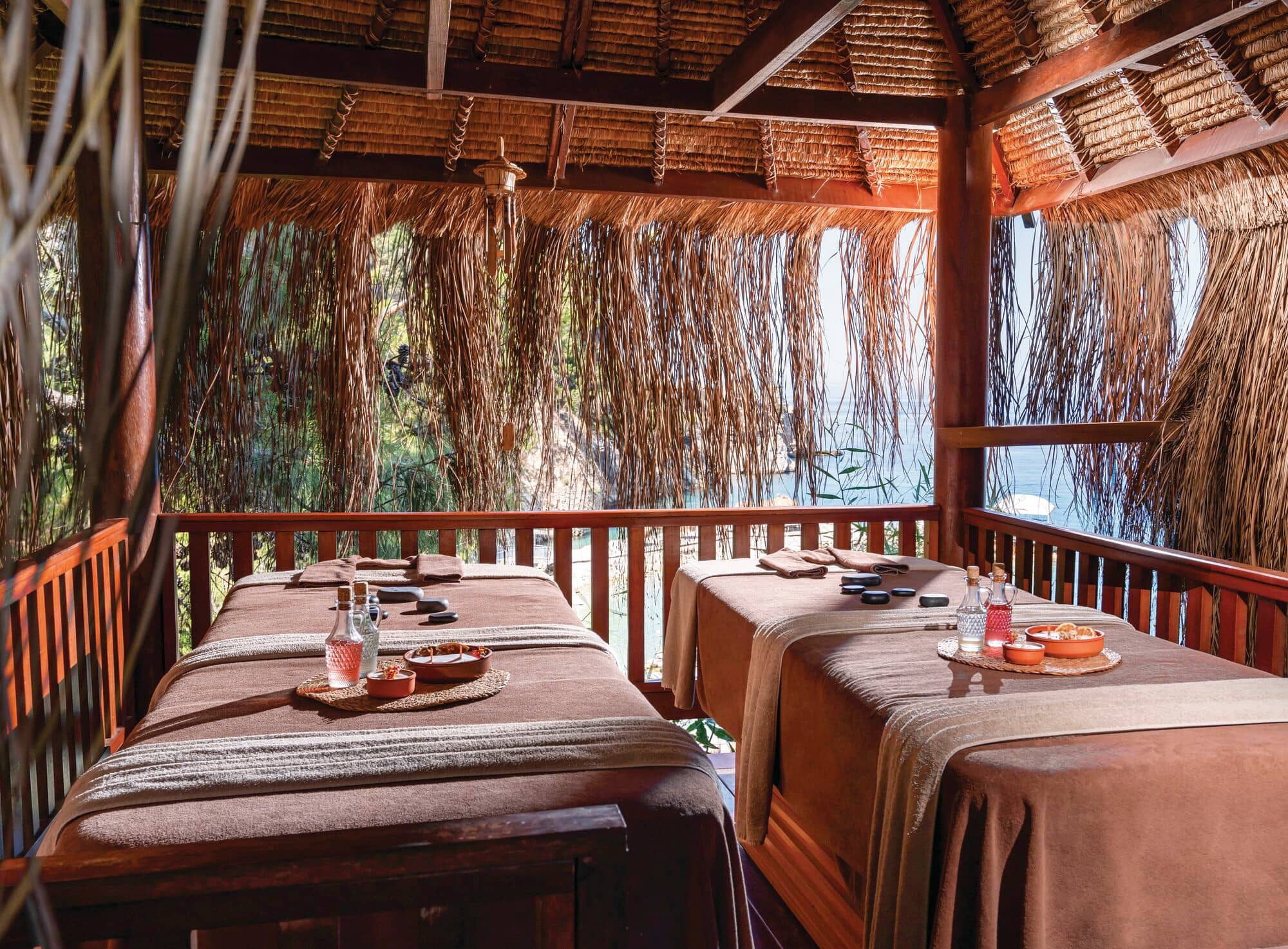 An ppen-air spa treatment pavilion with two massage beds set beneath a rustic wooden structure with a thatched roof.