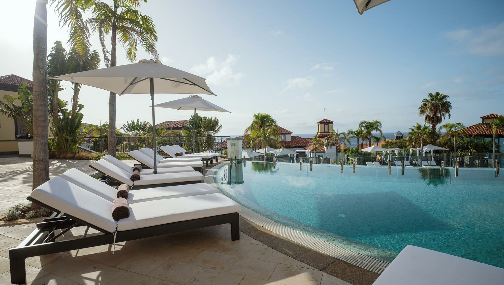 A luxurious poolside area with white cushioned lounge chairs, umbrellas, and an infinity pool surrounded by palm trees.