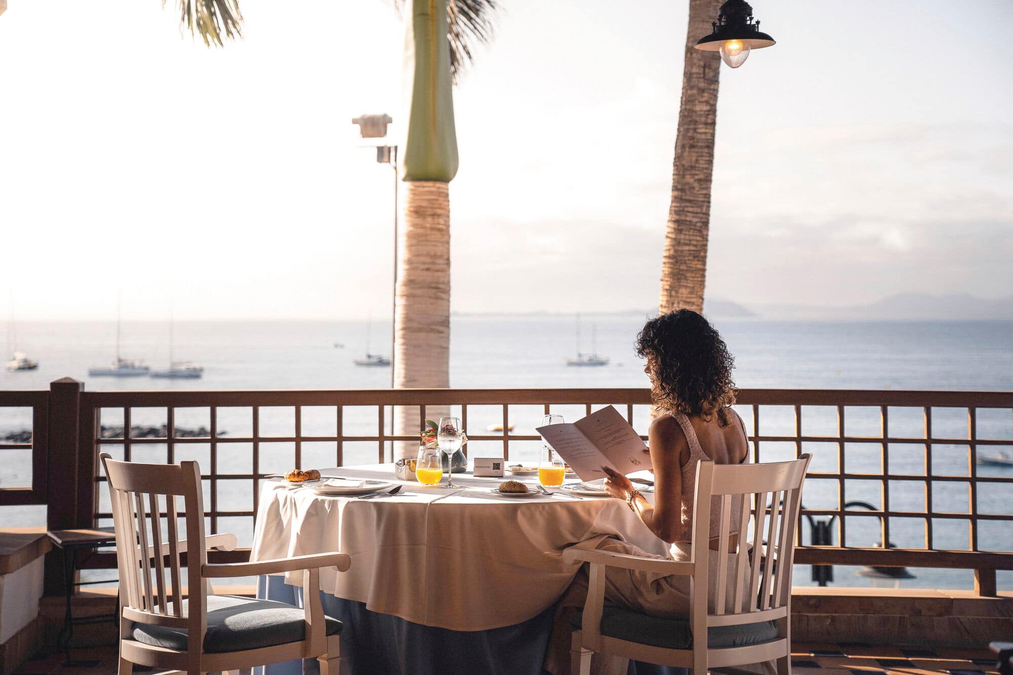 A person reading a book at a dining table with a sea view.