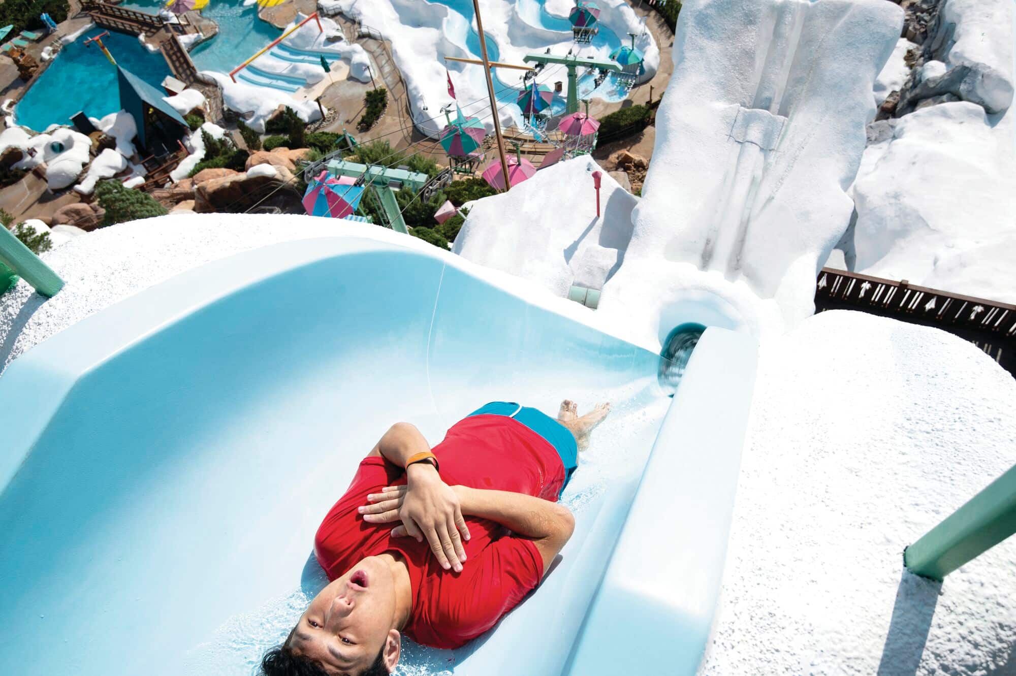 A person wearing a red shirt and blue shorts slides down a steep, light blue water slide with arms crossed and eyes closed.