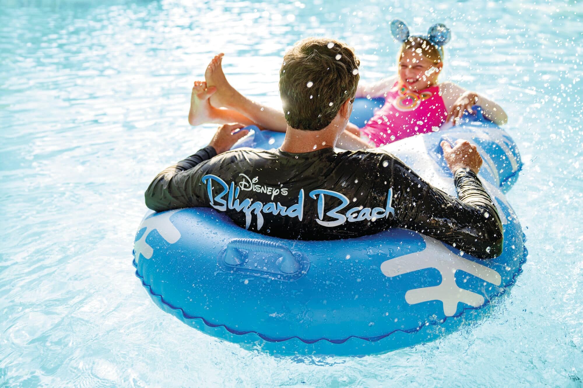 Two people enjoying water ride at Disney’s Blizzard beach. One wears a black long-sleeve shirt with the park's name, and the other wears a pink swimsuit with mouse ears. They sit in a blue inflatable tube surrounded by splashing water.