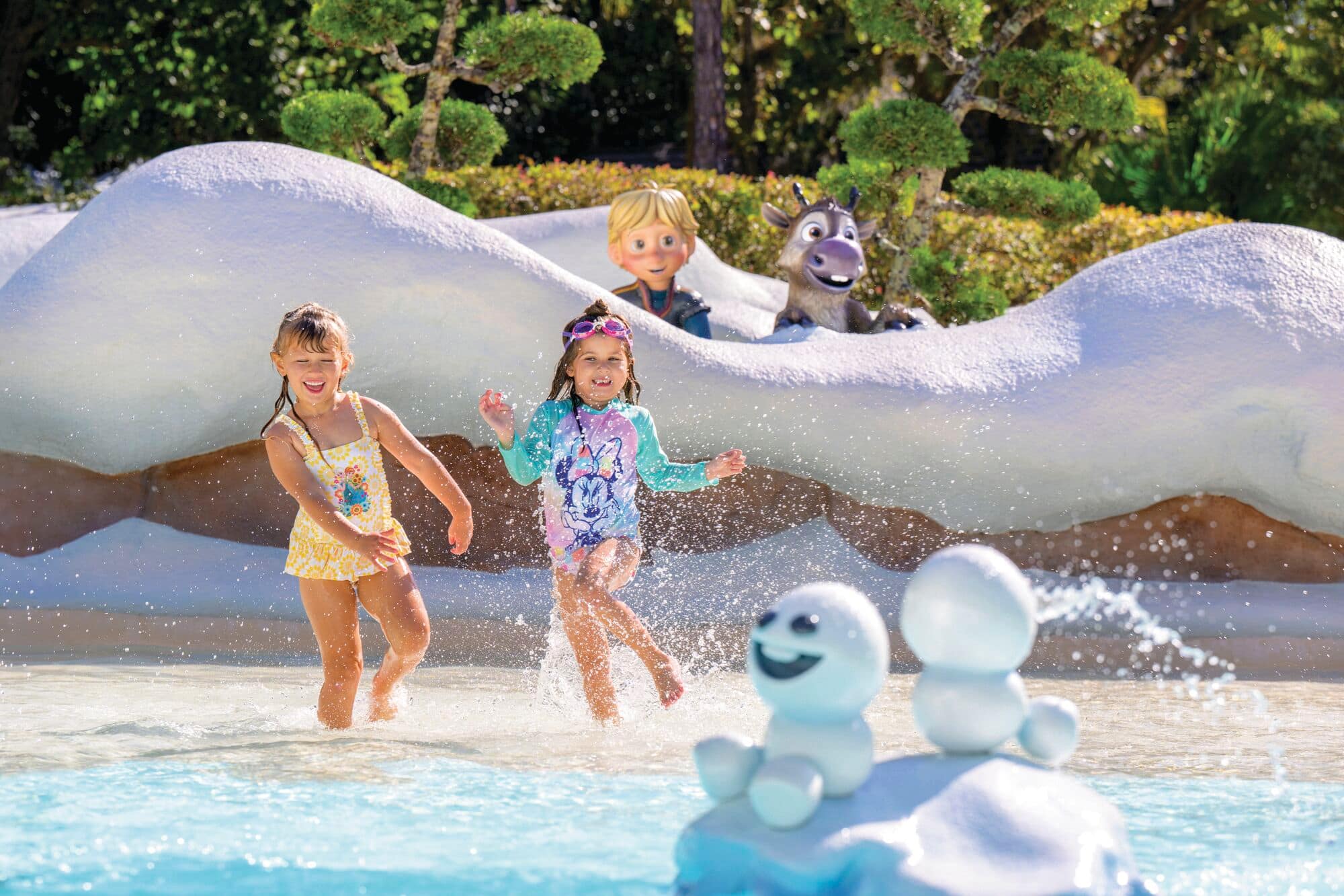 Two children play in a shallow water area with splashes around them. Behind them are snow-like decorations and characters from Disney's 'Frozen,' including Kristoff, Sven, and two small snowmen. The scene is bright and sunny, evoking a playful atmosphere.