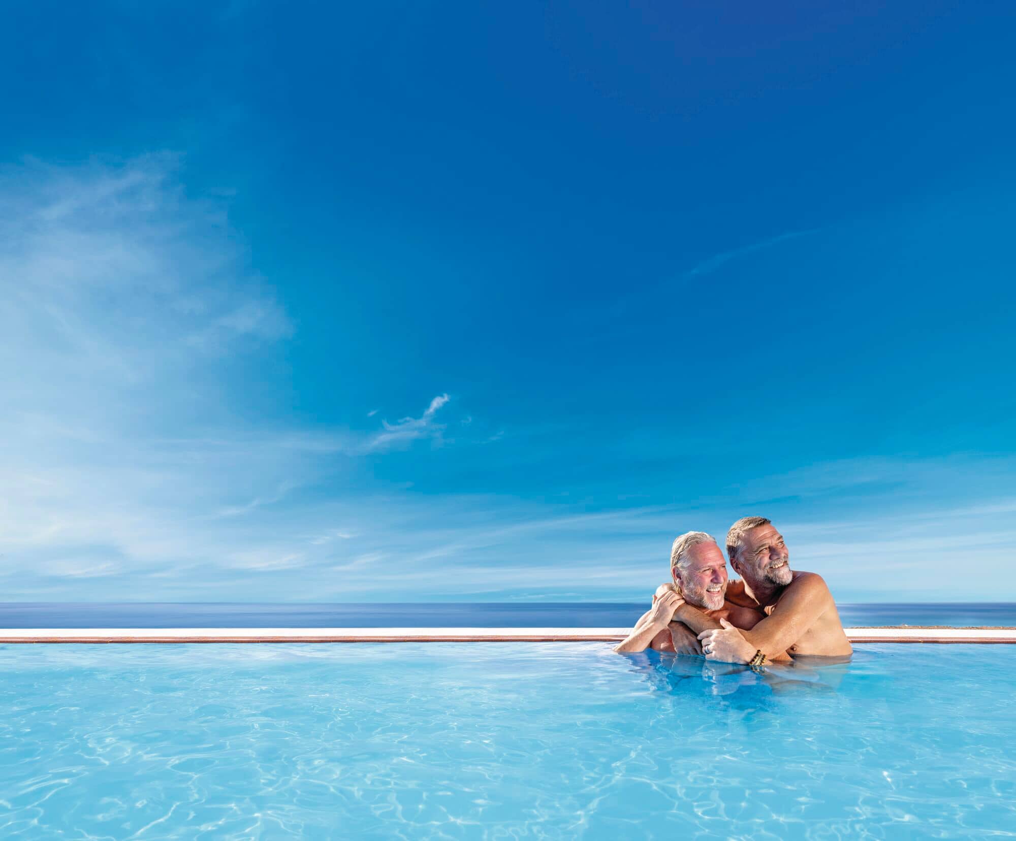 Two smiling men embrace in a turquoise infinity pool overlooking the ocean under a bright blue sky with wispy clouds.