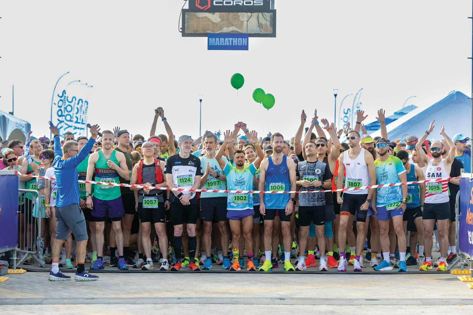 Marathon runners gathered at the starting line behind red tape, arms raised in anticipation, with green balloons overhead and Marathon signage visible.