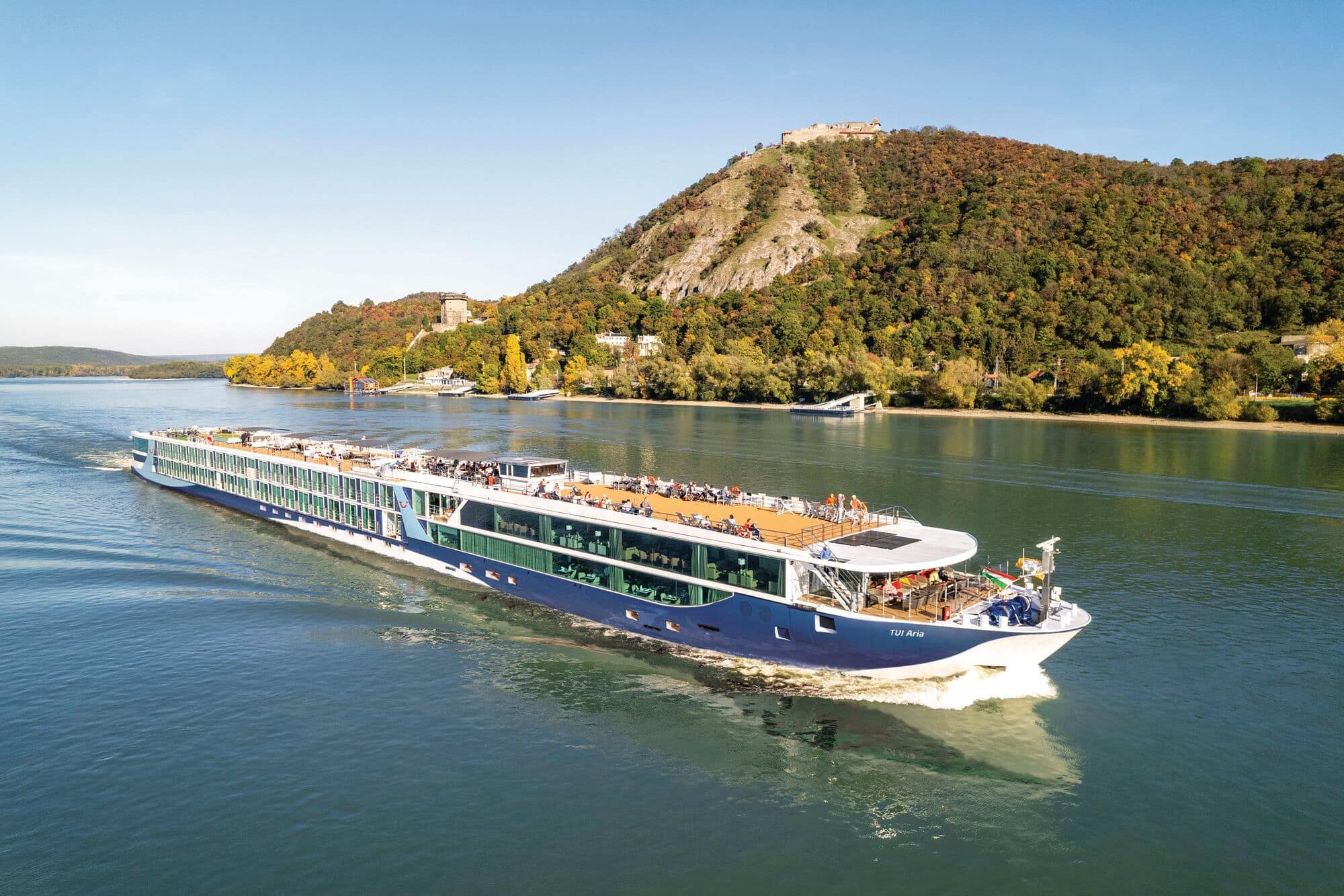 Exterior of the TUI Aria cruising on a river with green hilly banks.