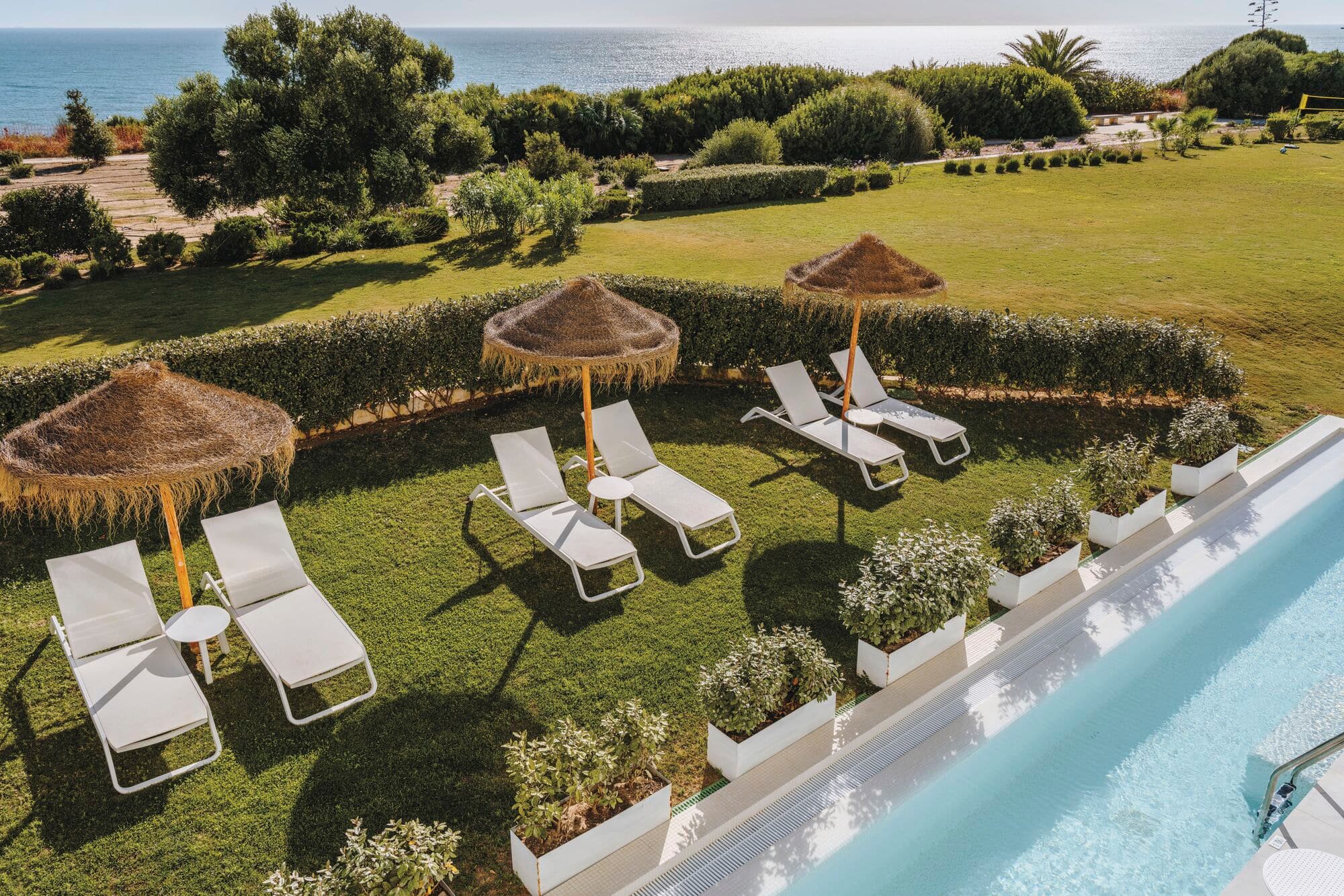 Elevated view of resort pool area with bright turquoise swimming pool on right, six white sun loungers arranged in rows on manicured lawn under three thatched umbrellas.