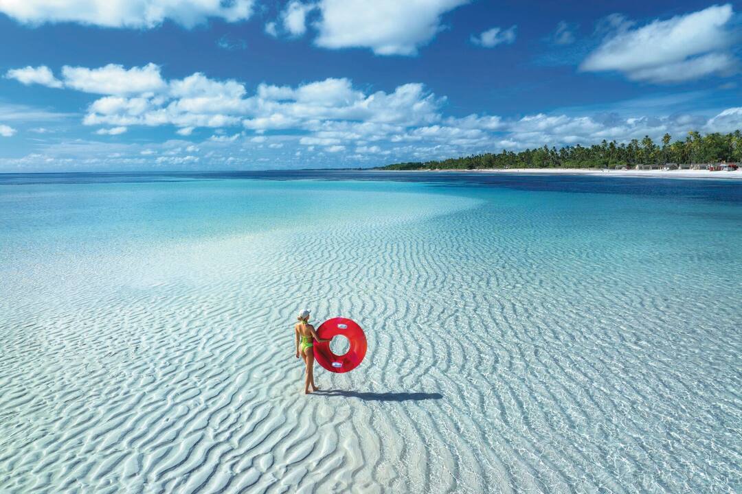 A woman stands on a vast swathe of pale sand, covered by crystal blue waters