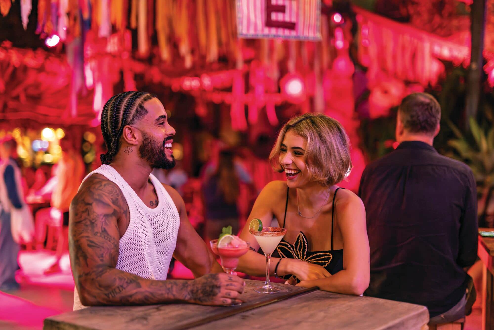 Two people smiling and conversing over cocktails at a wooden table in a tropical bar with pink lighting.