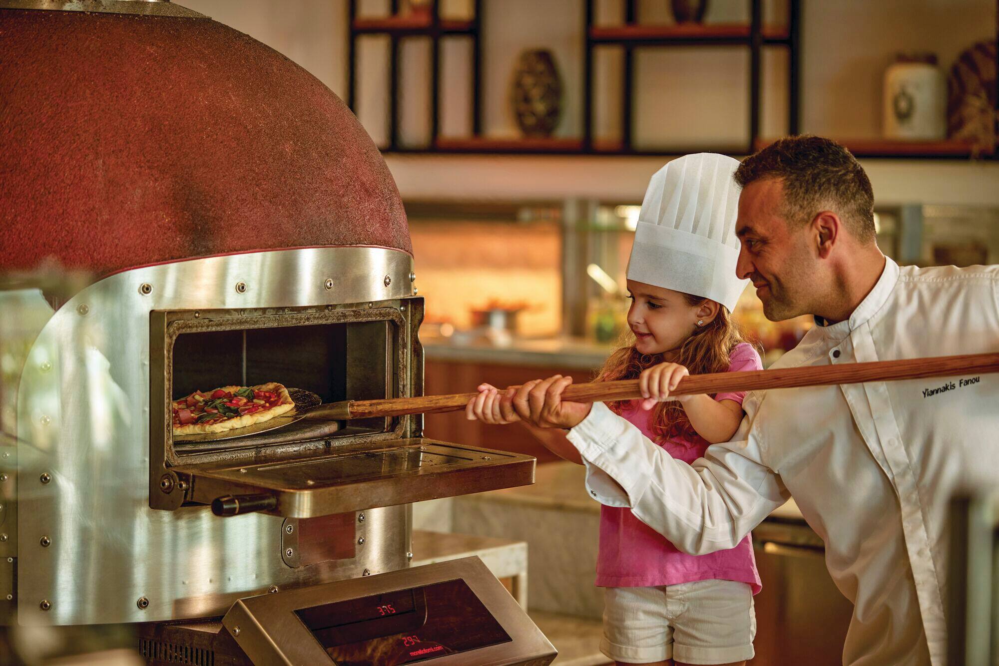 A child helping a chef to put a pizza into a wood fire oven.