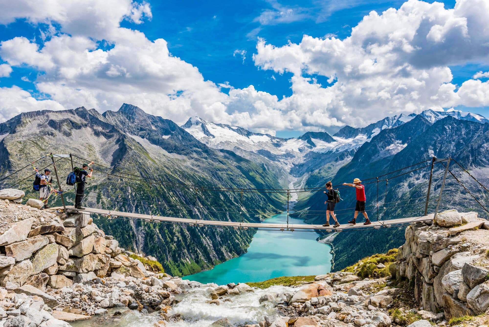 Two walkers crossing a suspension bridge with mountains and a blue lake in the background. 