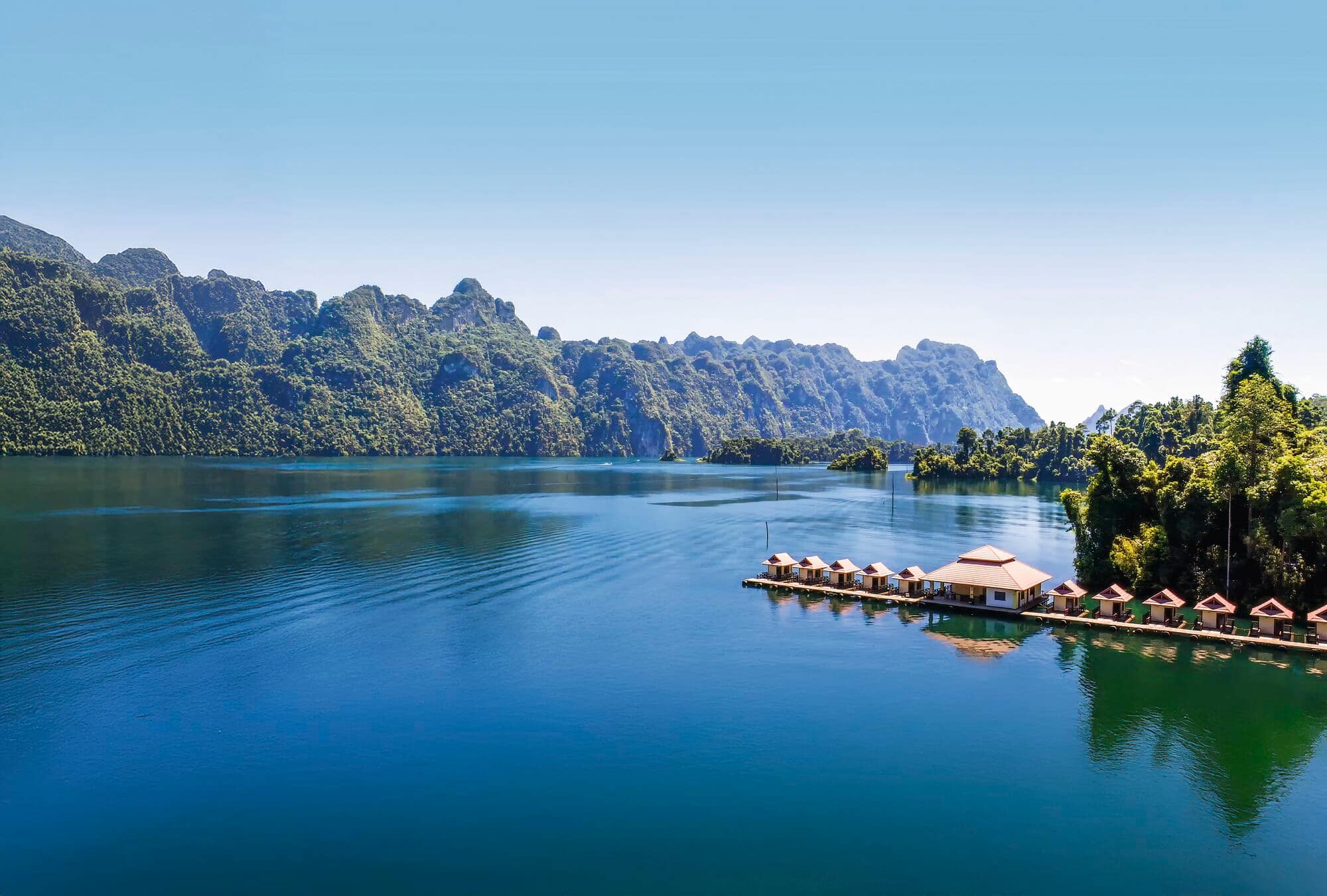 A lake surrounded by limestone mountains covered in green rainforest. A row of floating bungalows sits along the shoreline.