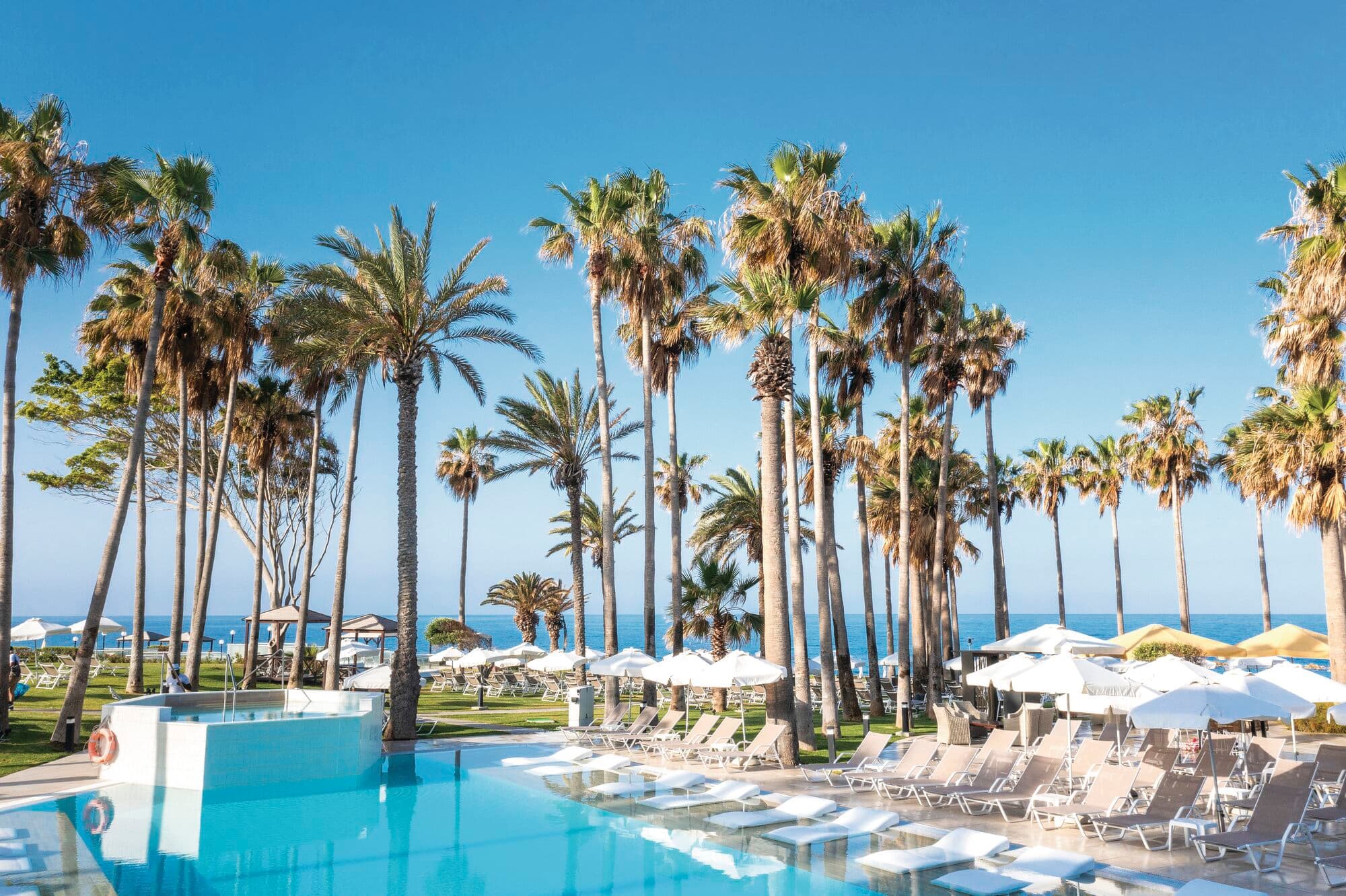 Infinity-style outdoor swimming pool with submerged loungers, surrounded by rows of beige sunbeds and white umbrellas. Tall palm trees frame the pool area, with a clear blue sky and ocean visible in the background.