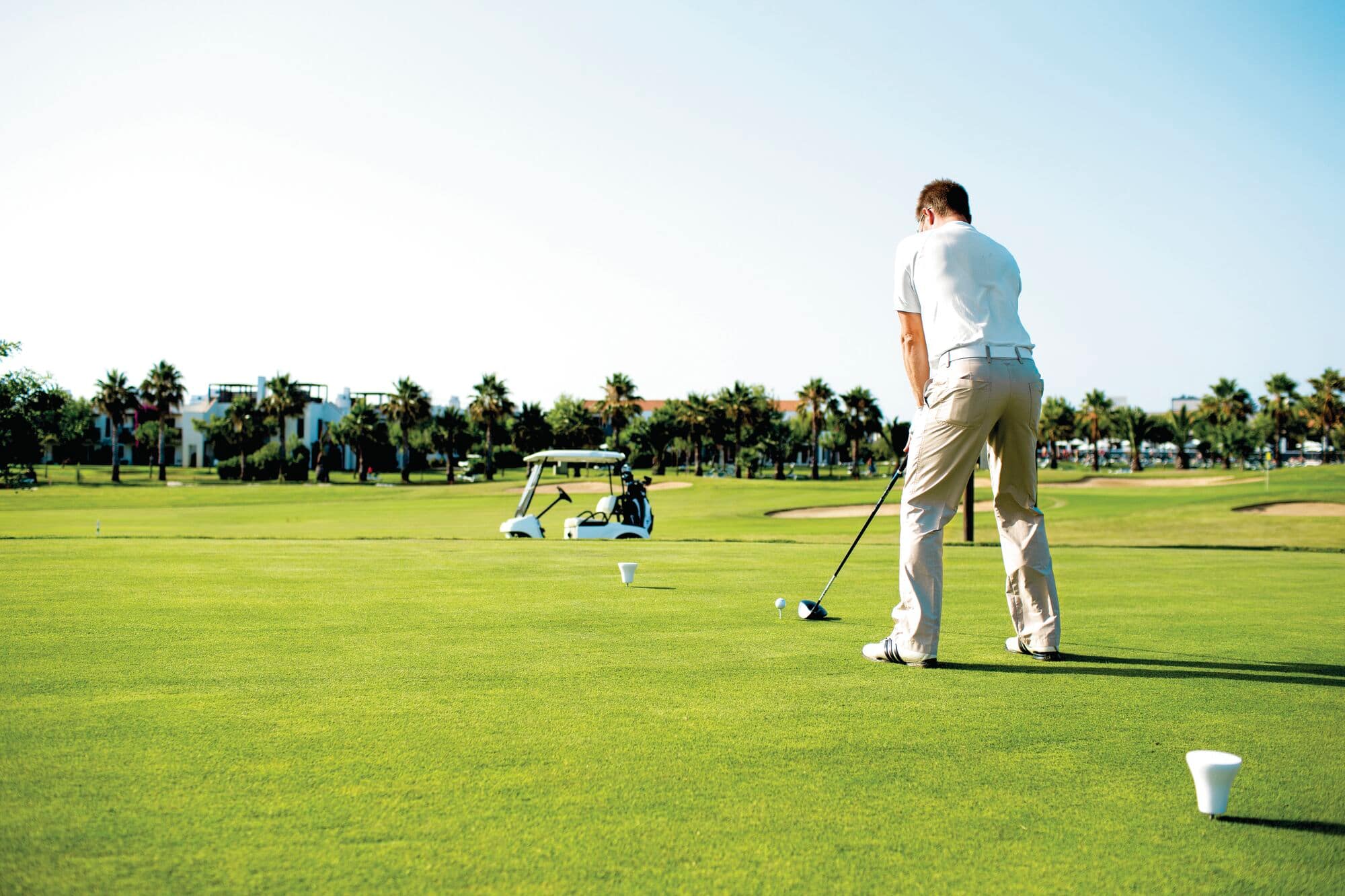 Golfer in light polo and khaki pants preparing to tee off on a tropical golf course with palm trees and resort buildings in the background.