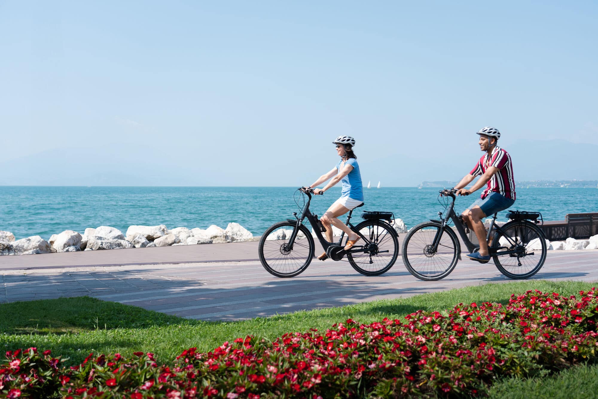 Two people cycling next to other on a path beside a lake. 