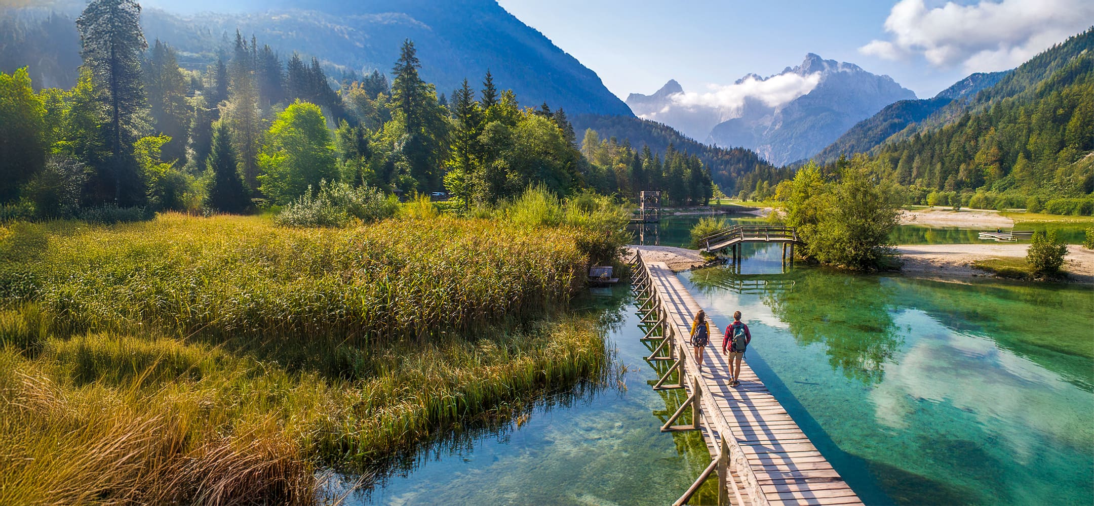Two people walking across a boardwalk over a small lake with mountains in the background.