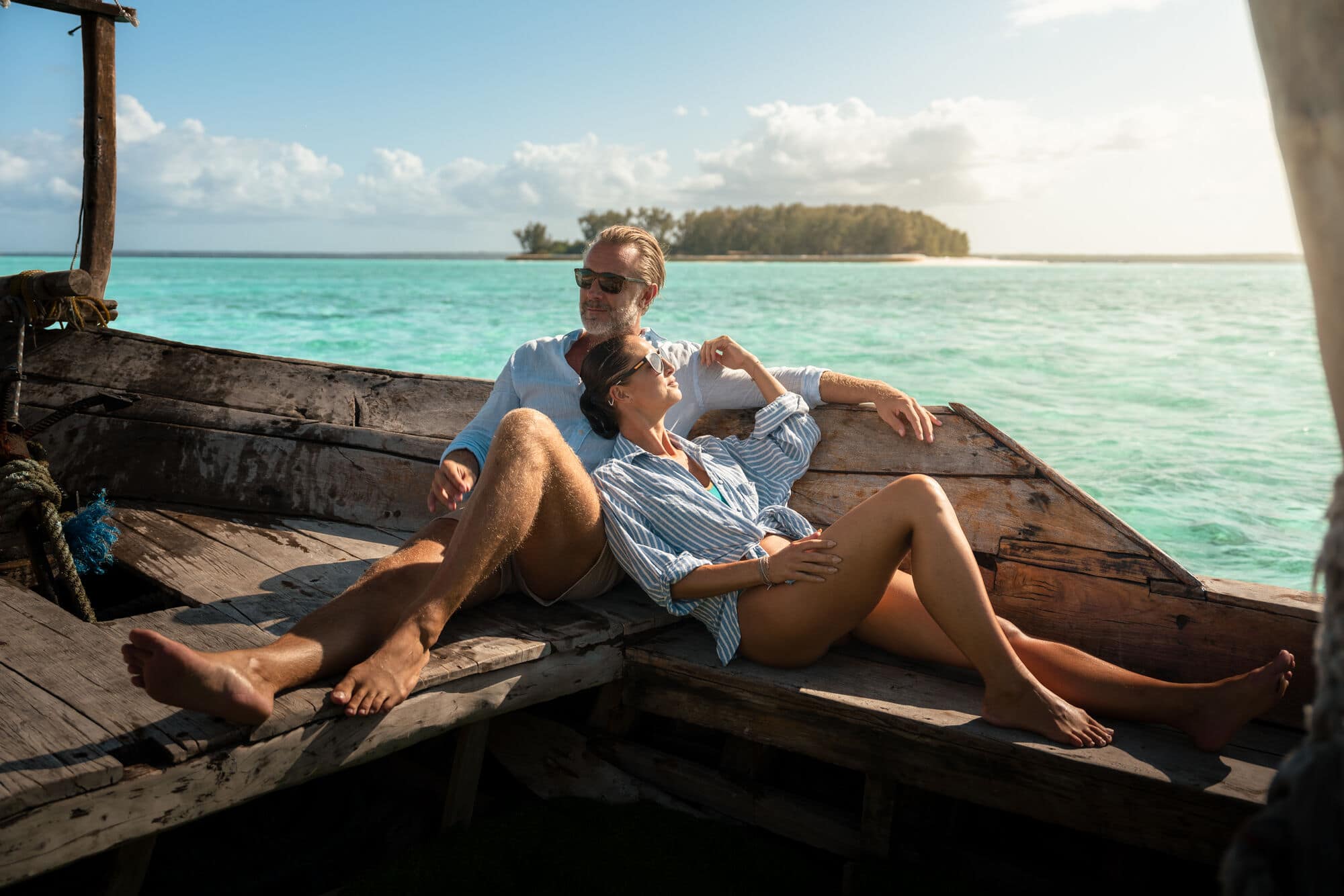 A couple relax together on a wooden boat with a small island in the background.