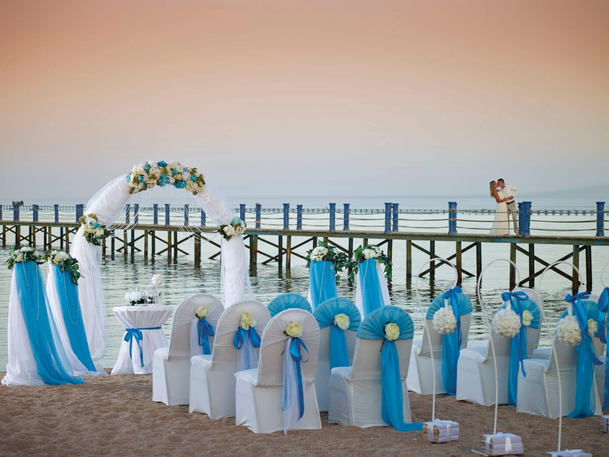 A couple kiss on a pier overlooking a romantic beach wedding ceremony set-up.
