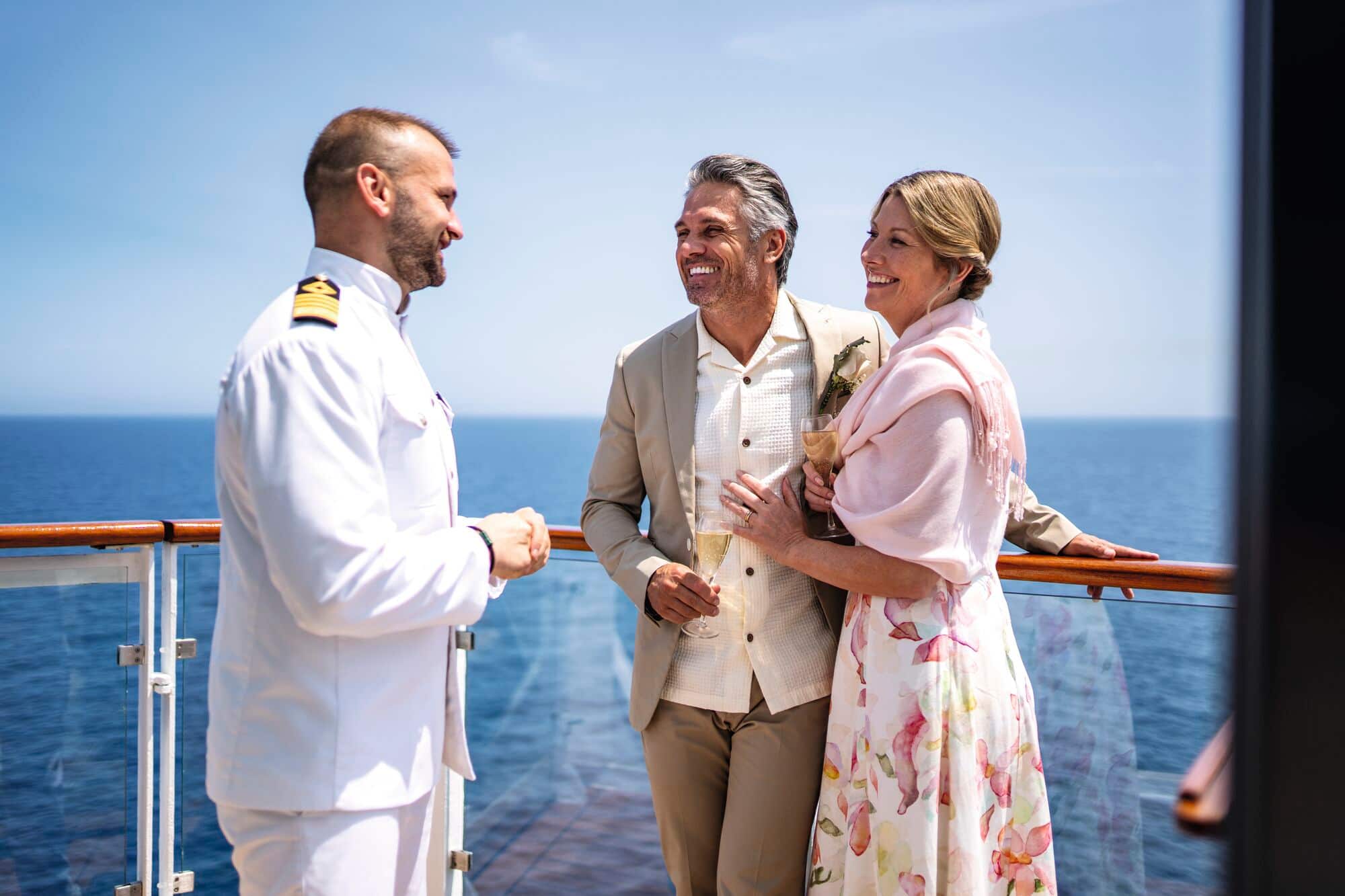 A cruise ship captain in a white naval uniform speaking with a couple on the deck of the ship.