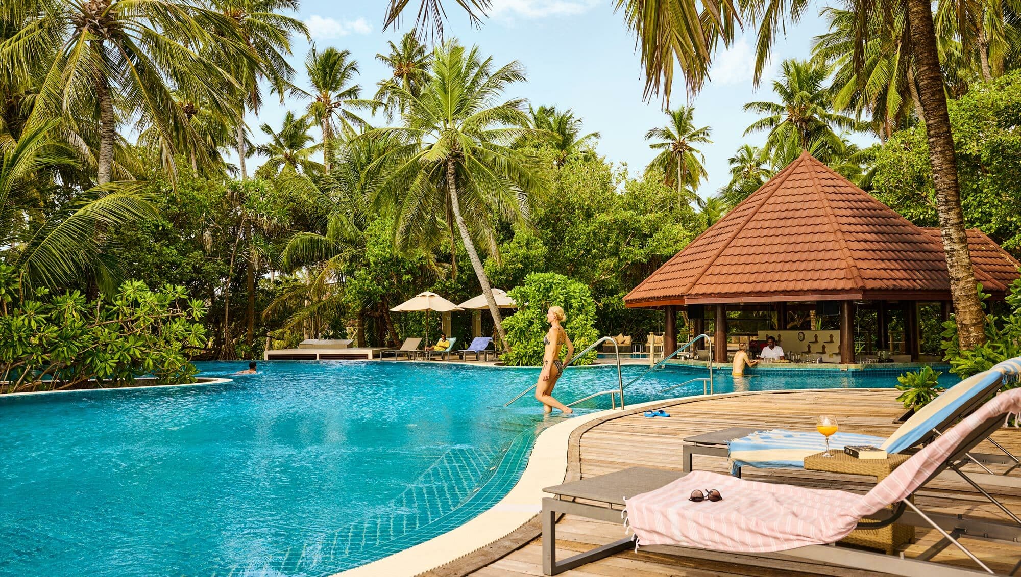 Tropical resort swimming pool surrounded by palm trees with sun loungers.