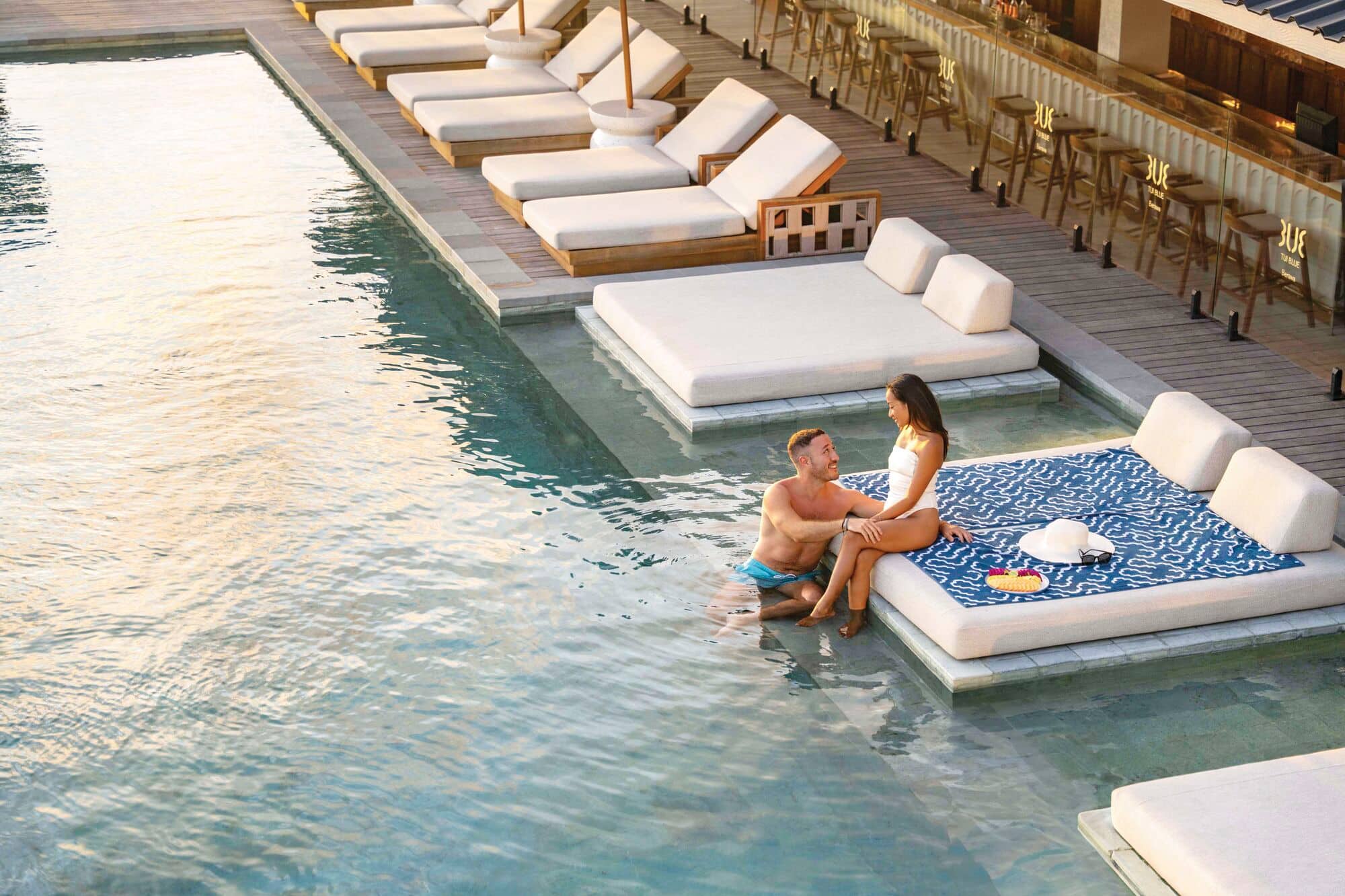 A couple sitting on the edge of an in-water lounger in a hotel swimming pool.