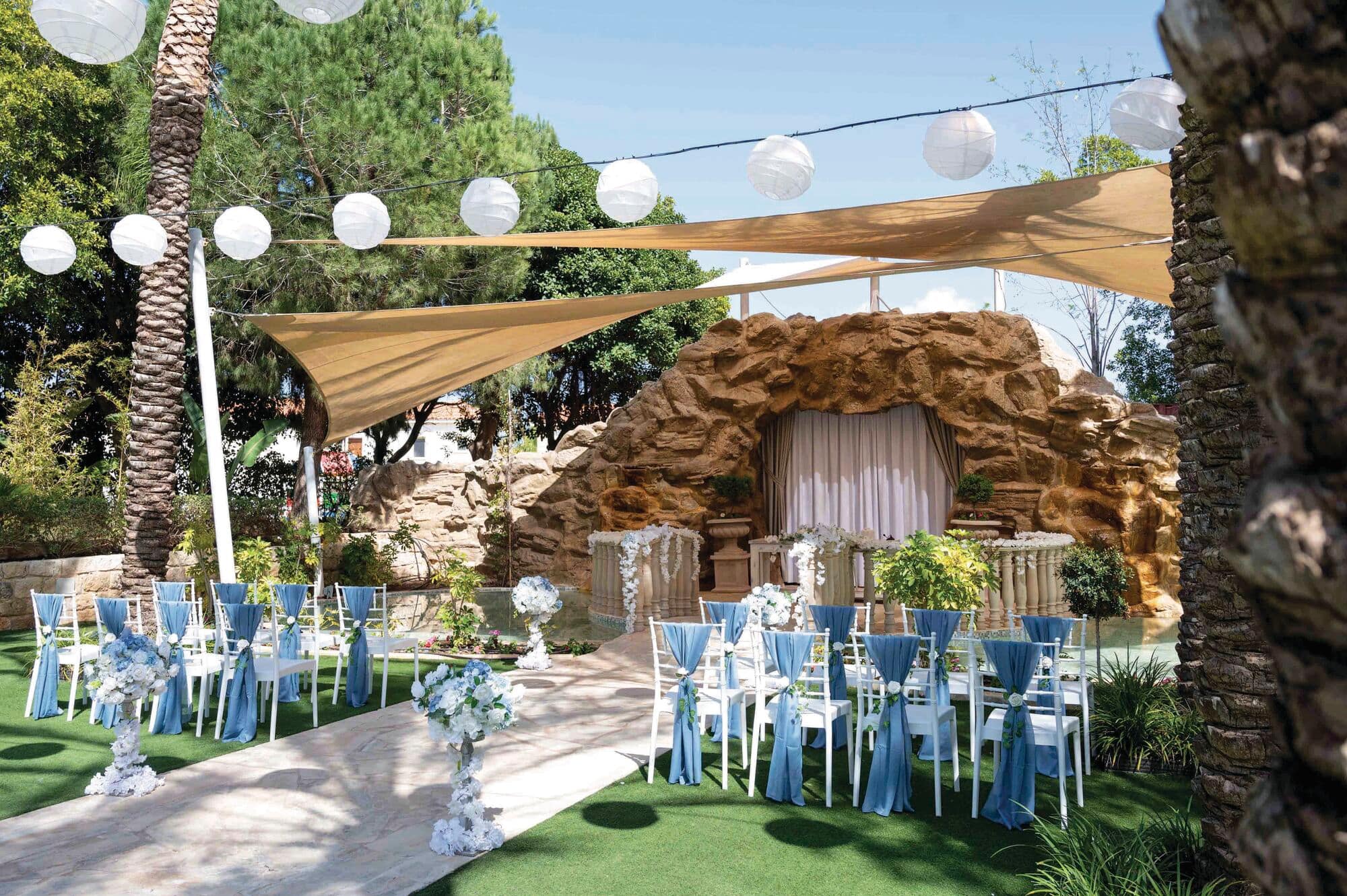 A wedding setup sits before a grotto in this hotel’s gardens. Neutral canopies and large lights hang above rows of seats adorned with blue fabric.