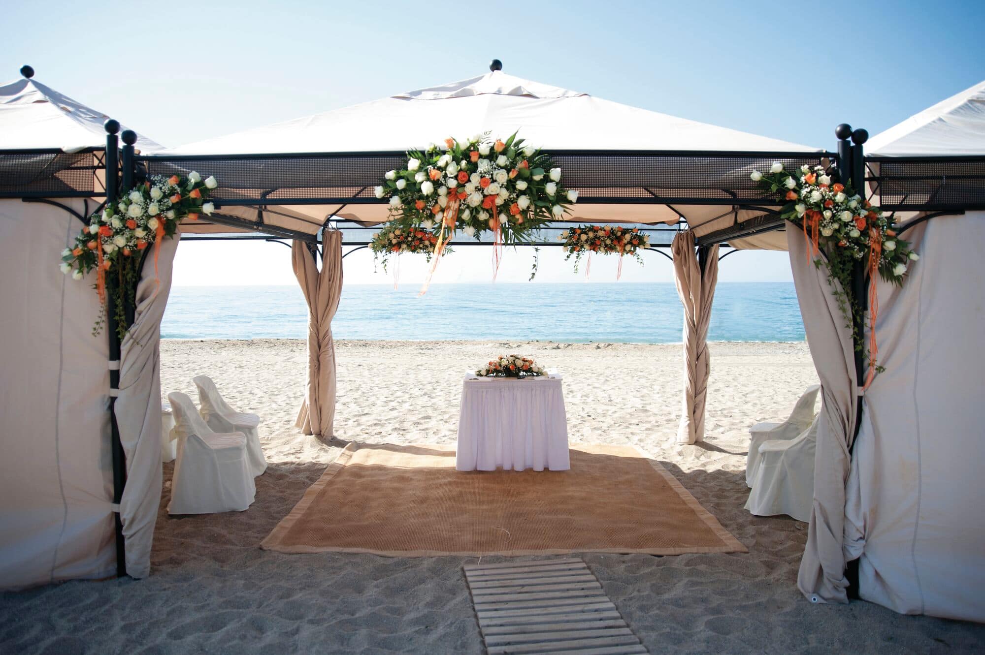 A beach wedding set-up under two white canopy tents decorated with orange and white floral arrangements.