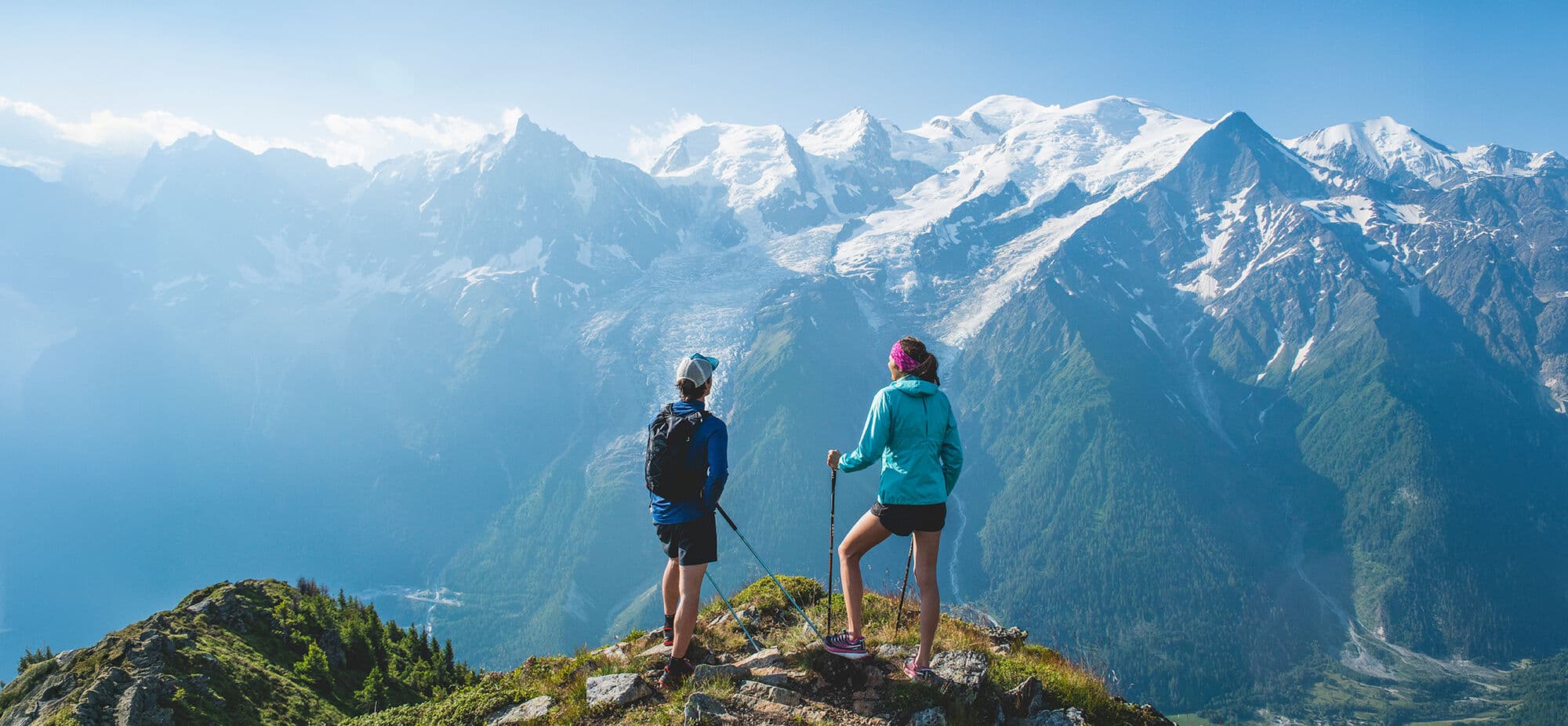 Two hikers standing on a summit overlooking a snowy mountain range including Mont Blanc. 