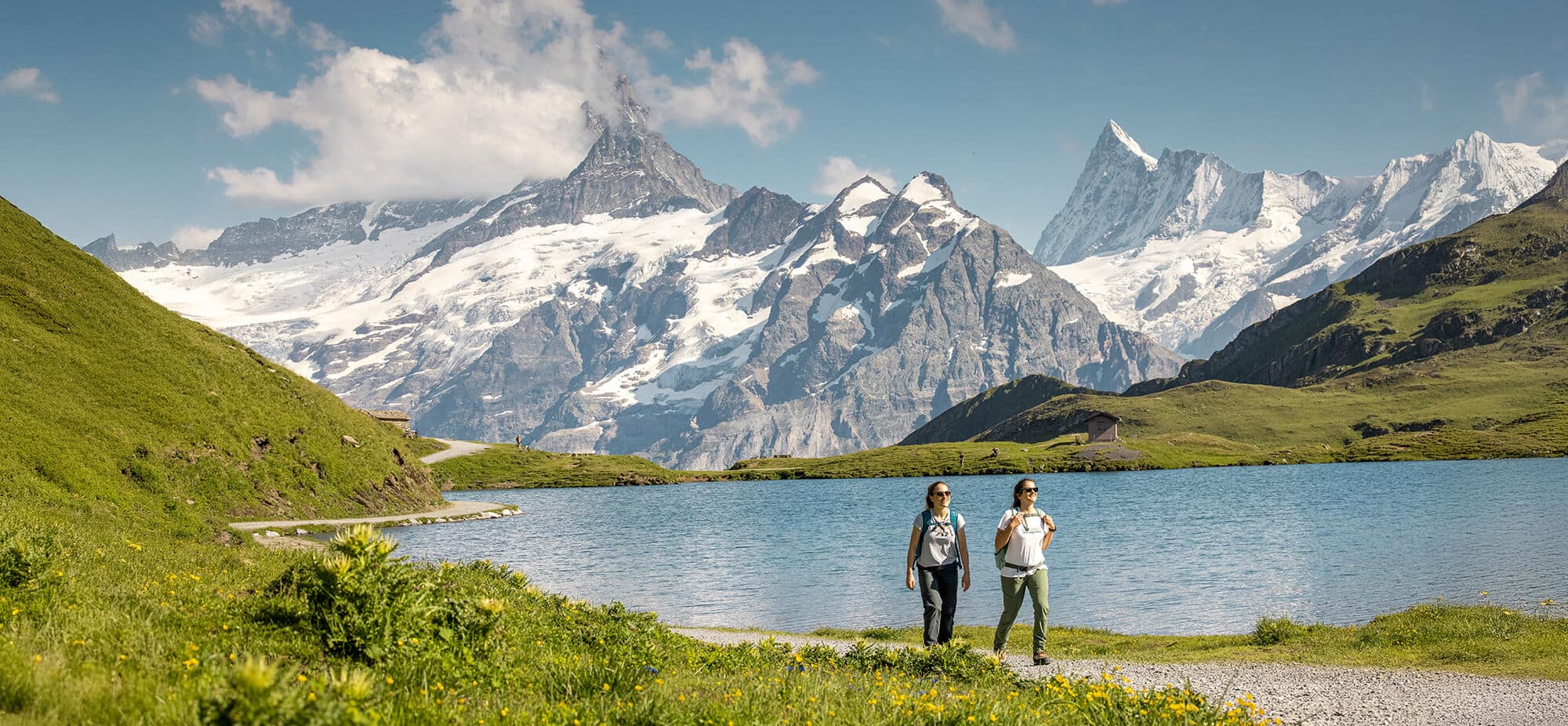 Two women walking alongside a lake near Grindelwald, with mountains in the background.