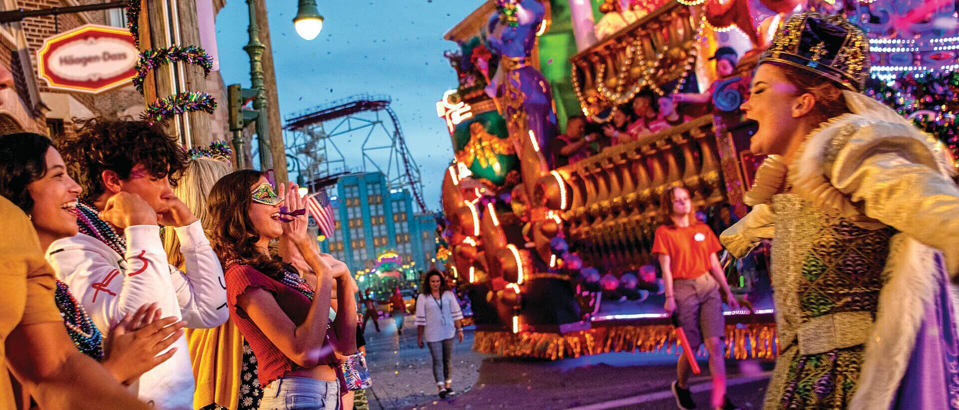 People in Mardi Gras costumes watching a singer in front of the park rides.