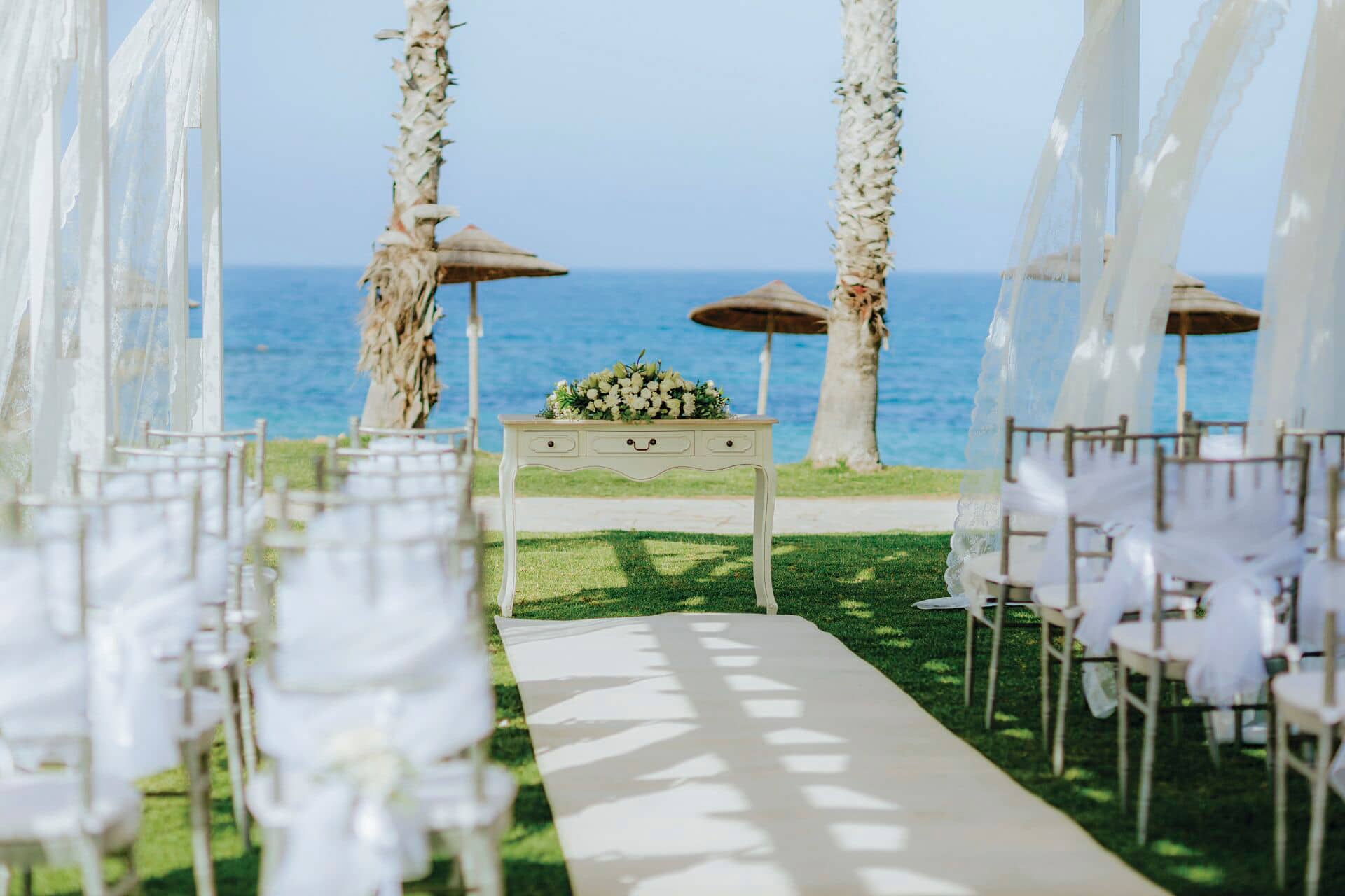 Looking down the aisle toward a simple, but elegant altar. A flower arrangement sits upon a table, with the sea and tropical parasols in the background.