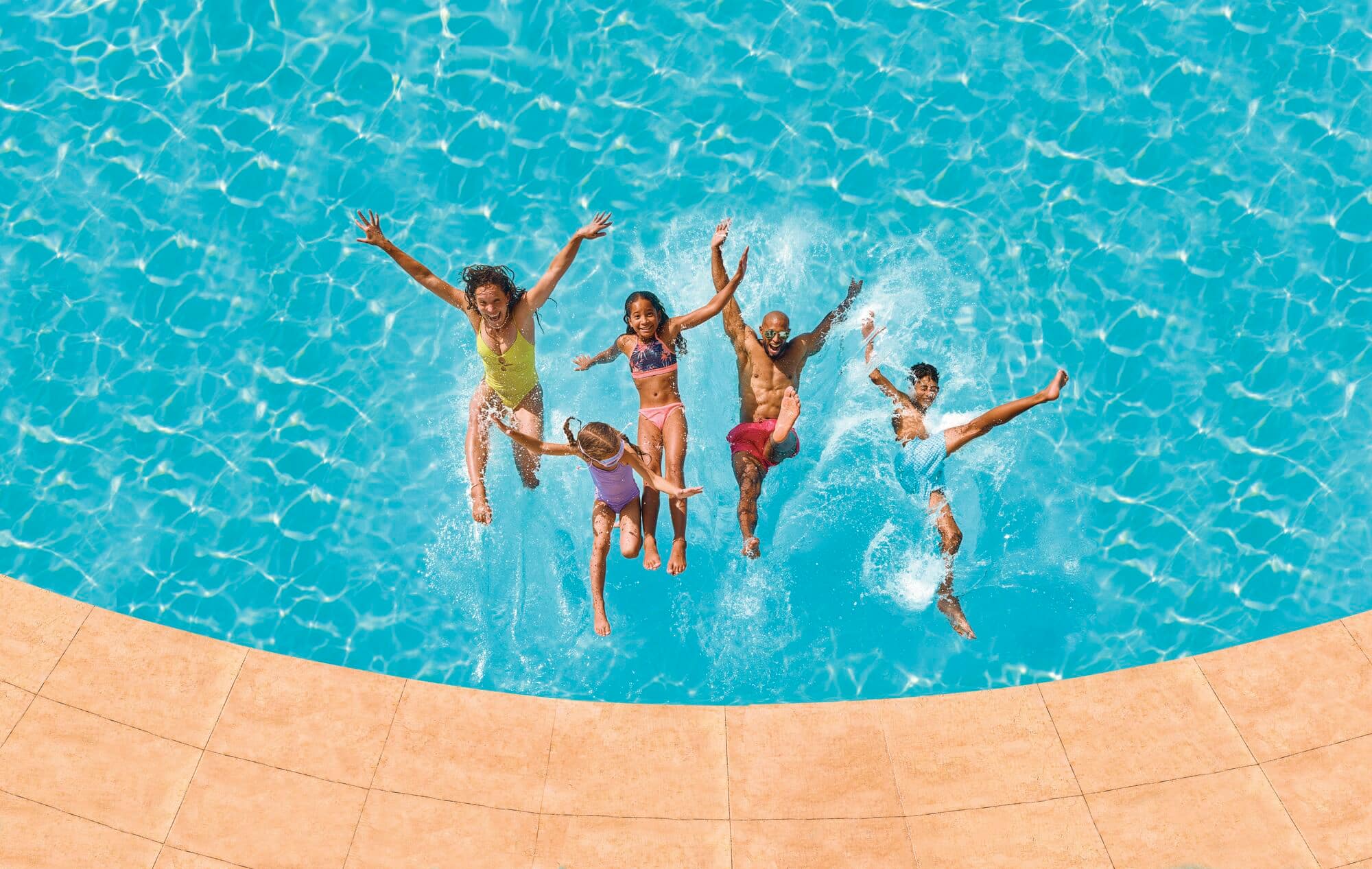Overhead view of five people jumping into a turquoise swimming pool simultaneously.