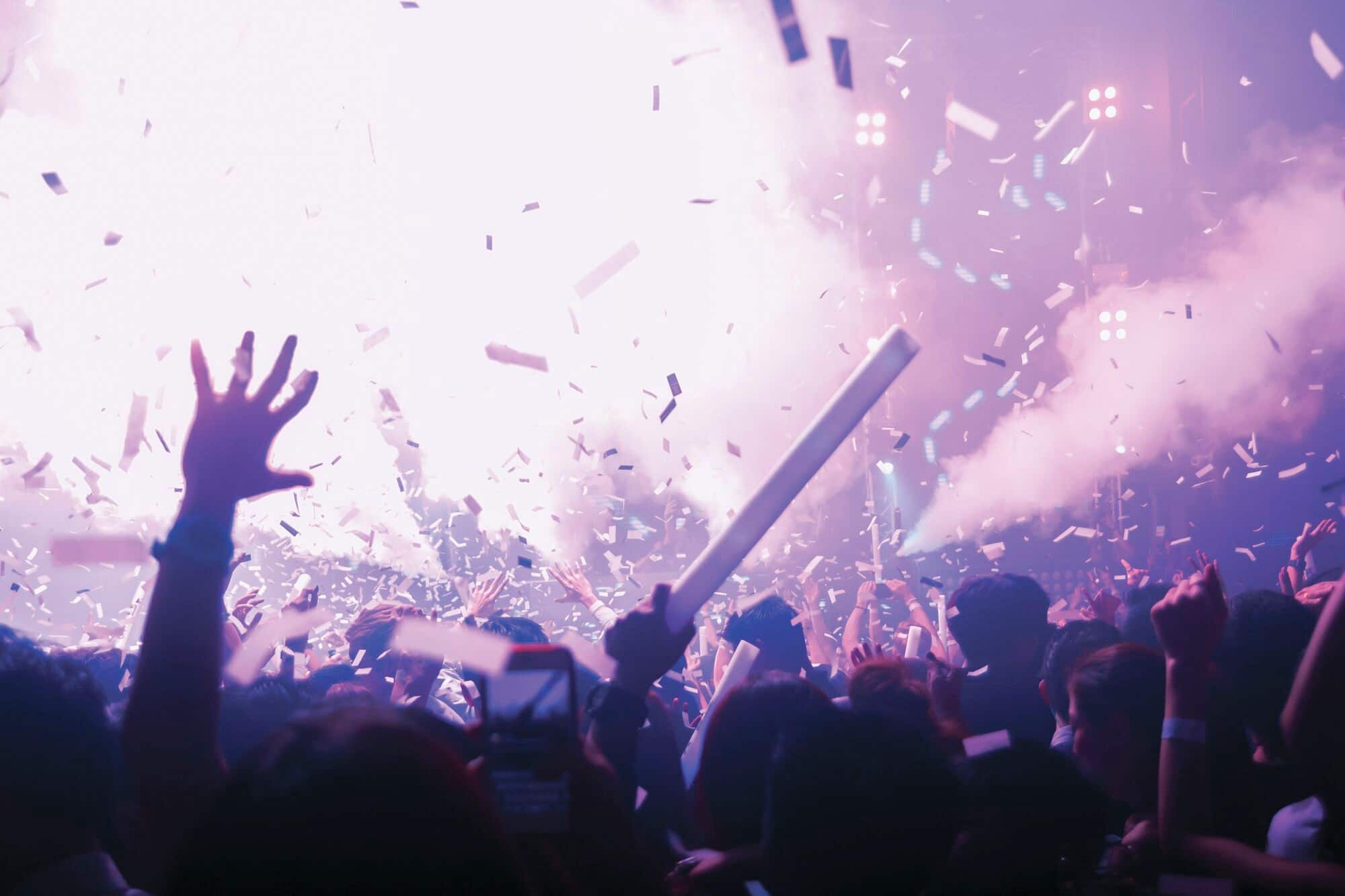 Crowd gathered at an outdoor event with confetti falling and stage lights visible in the background.