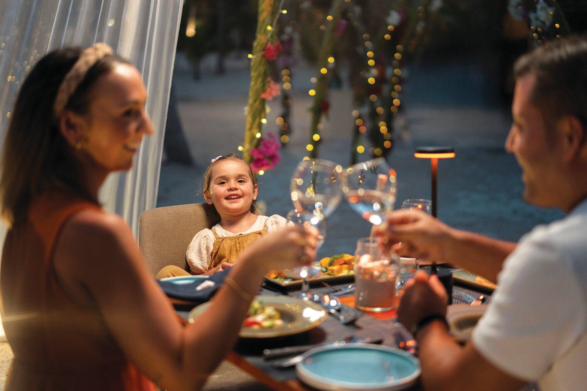 A smiling family toast glasses at a dining table surrounded by lights.