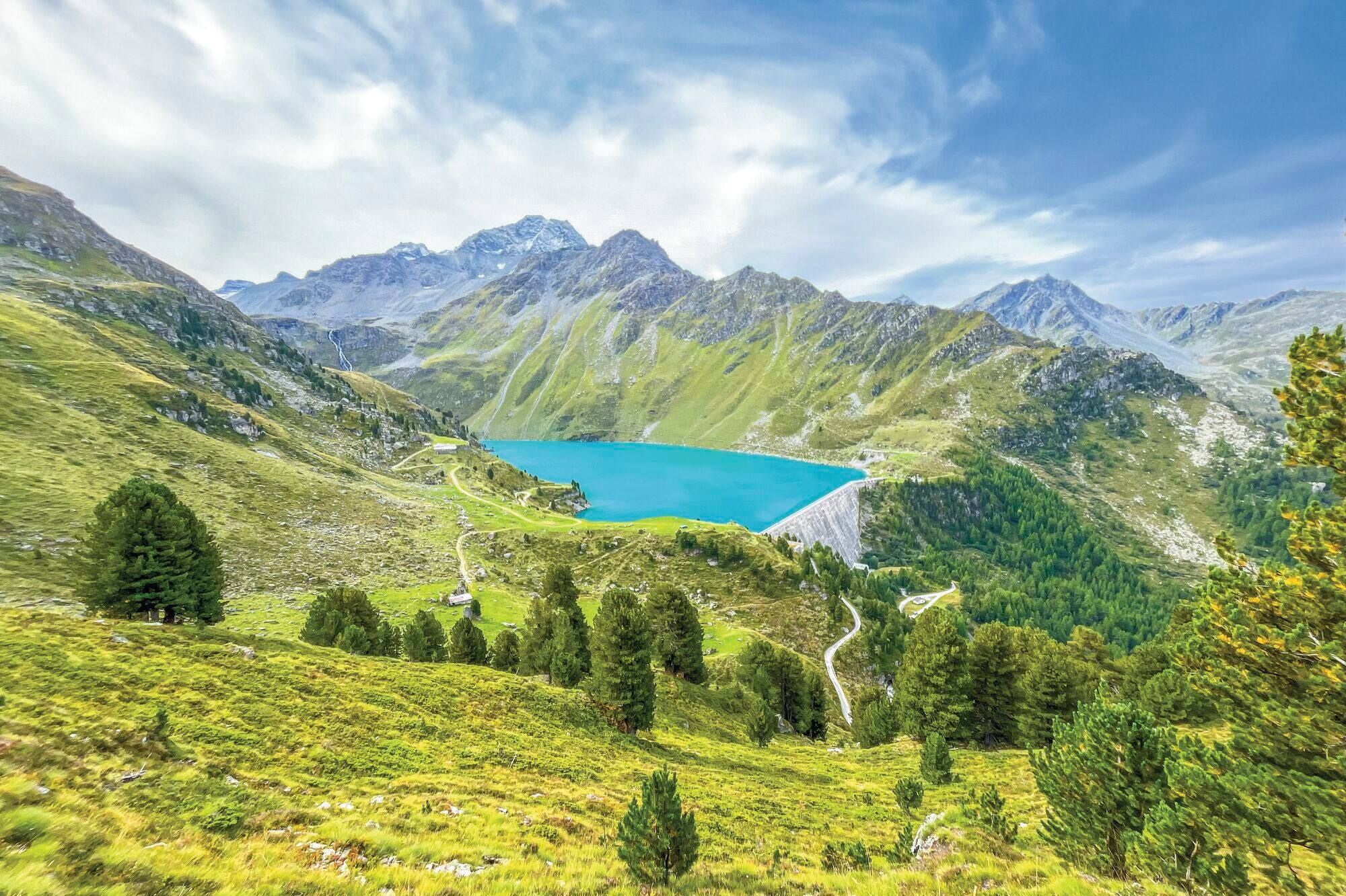 Turquoise reservoir nestled in Nendaz's alpine valley, surrounded by lush green meadows and snow-capped peaks under a blue sky.