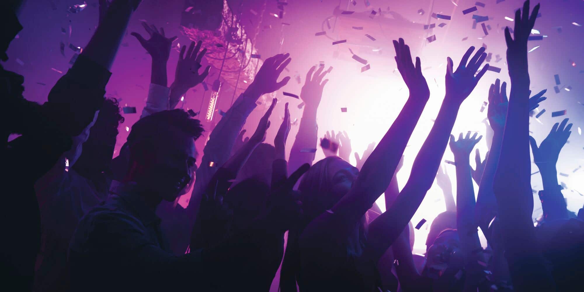 A crowd of people with their hands up in the air at a purple-lit nightclub.