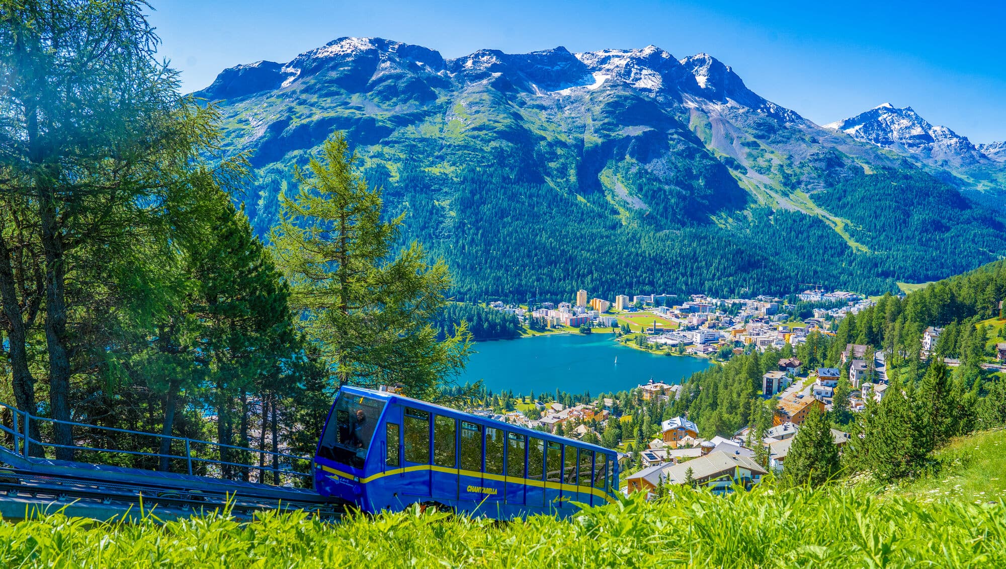 Wooden chalets with timber walls line a winding trail in a Swiss Alpine village, surrounded by towering mountains with forested slopes and snow-capped peaks.