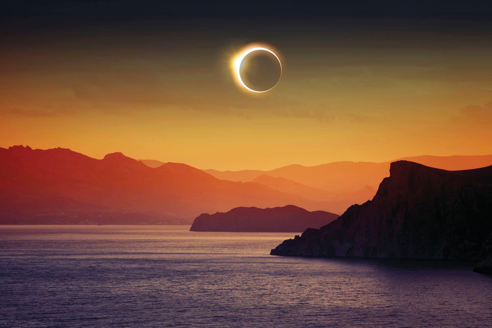 A solar eclipse above the sea, with mountains in the background.